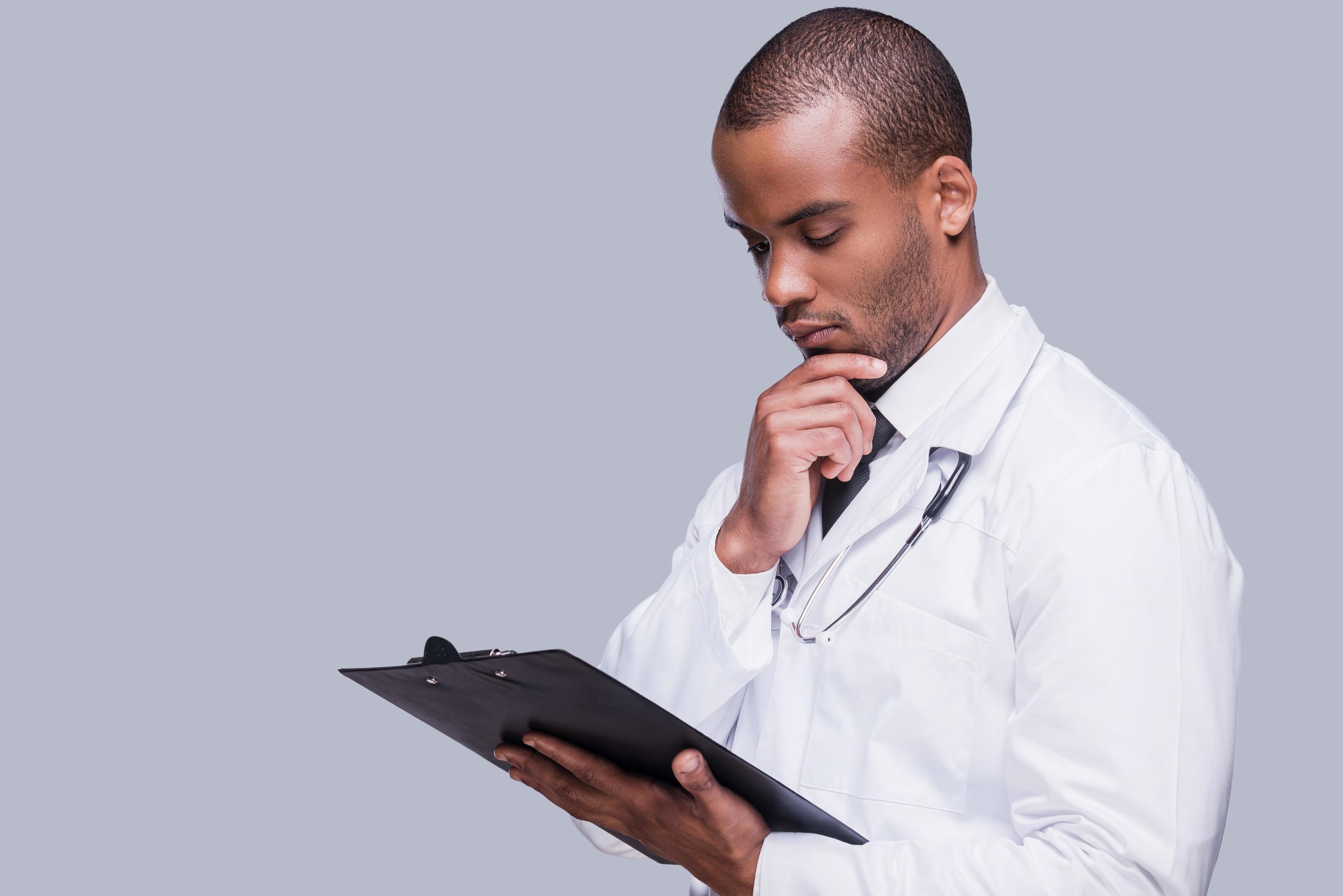 Thinking about proper medication. Thoughtful African doctor holding clipboard and looking at it while standing against grey background Stock Free