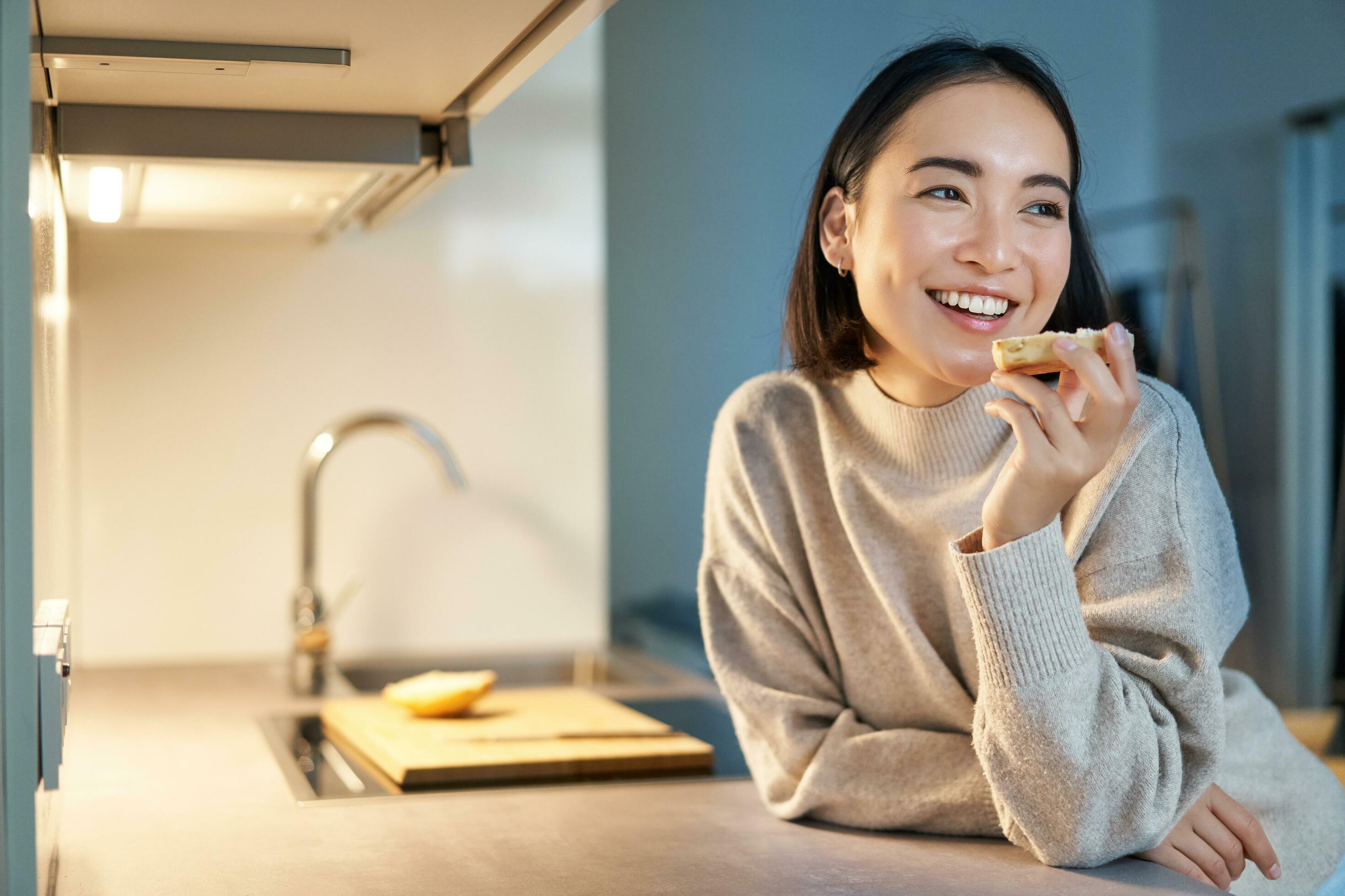 Portrait of smiling young happy woman staying at home, standing in kitchen and eating toast, looking aside Stock Free