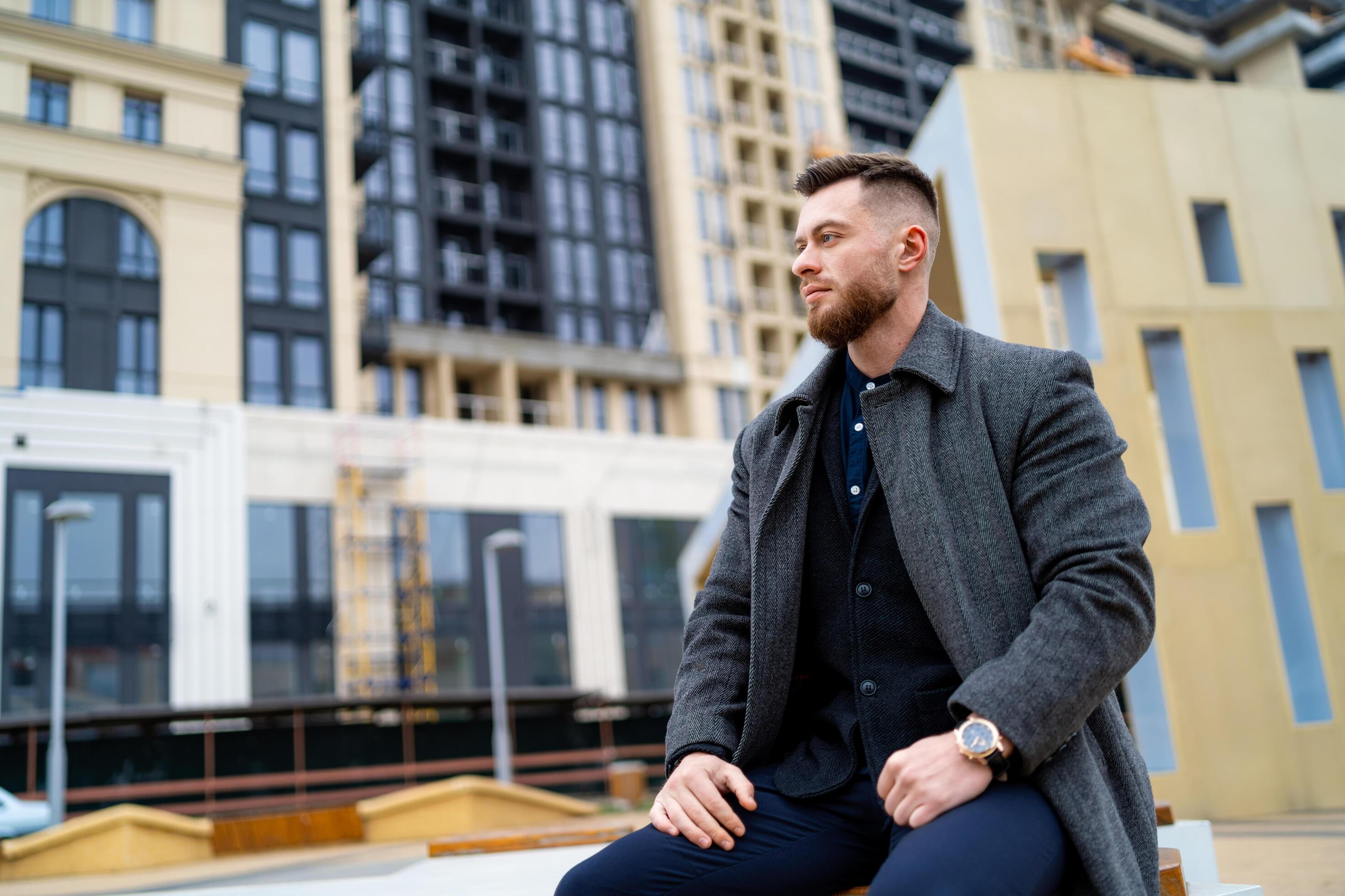 Man in formal clothes sitting on a street bench with hands on legs. Fashion and stylish handsome young male. Modern building on the background. Stock Free