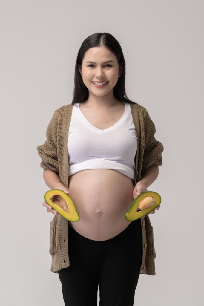 Portrait of Beautiful pregnant woman holding avocado over white background studio, health and maternity concept Stock Free