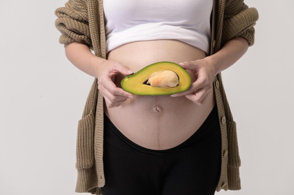 Portrait of Beautiful pregnant woman holding avocado over white background studio, health and maternity concept Stock Free