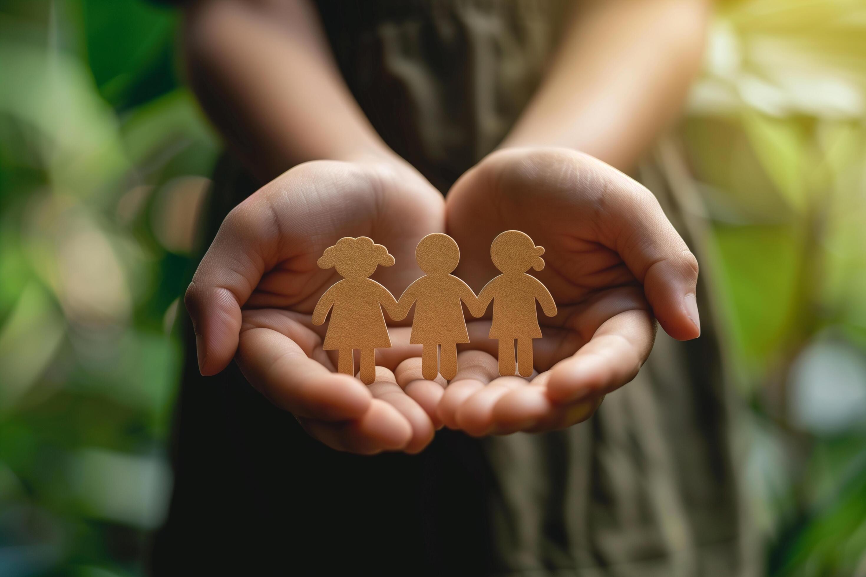 A hand holds a paper cutout of a family inside a heart-shaped house against a garden with sunlight. Stock Free