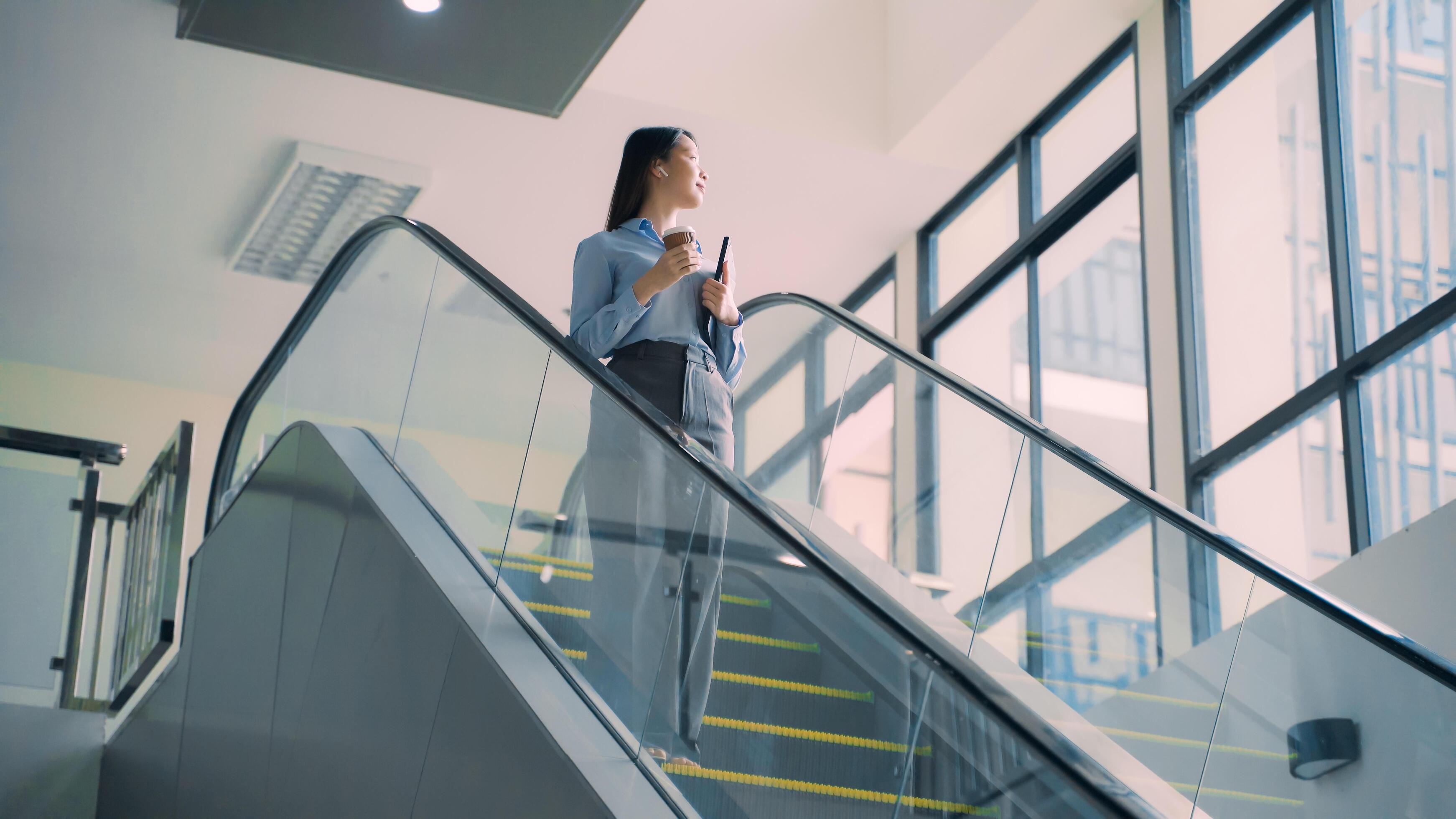 A business woman is walking down to escalator with a coffee cup in her hand Stock Free