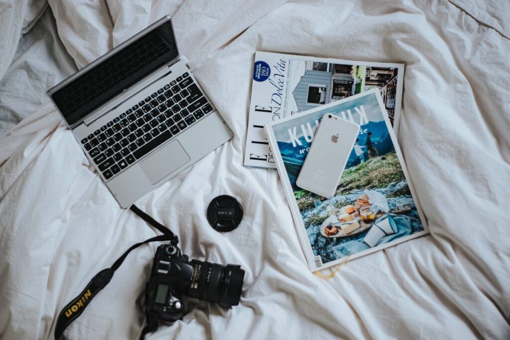 Silver laptop, a camera, magazines and other items on white bed sheets Stock Free