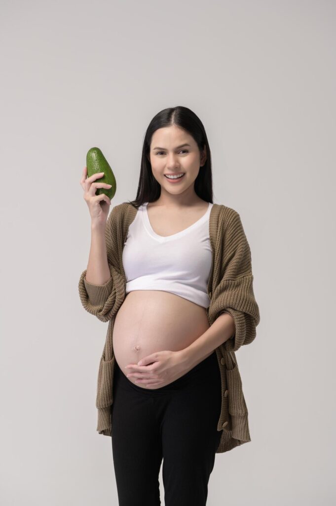 Portrait of Beautiful pregnant woman holding avocado over white background studio, health and maternity concept Stock Free