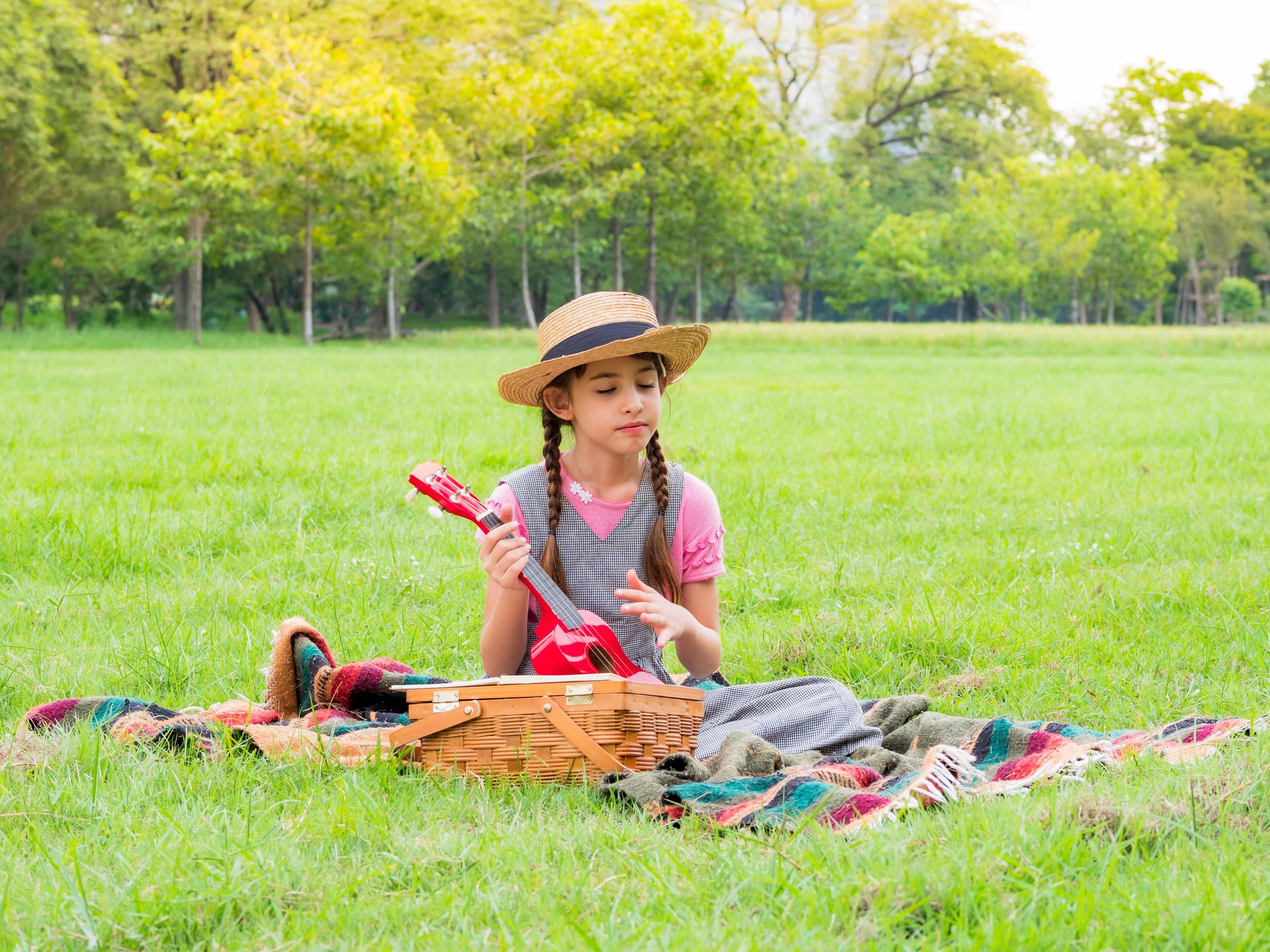 The girl sits on the grass and learns to play the ukulele, learning outside of school in the nature park Stock Free