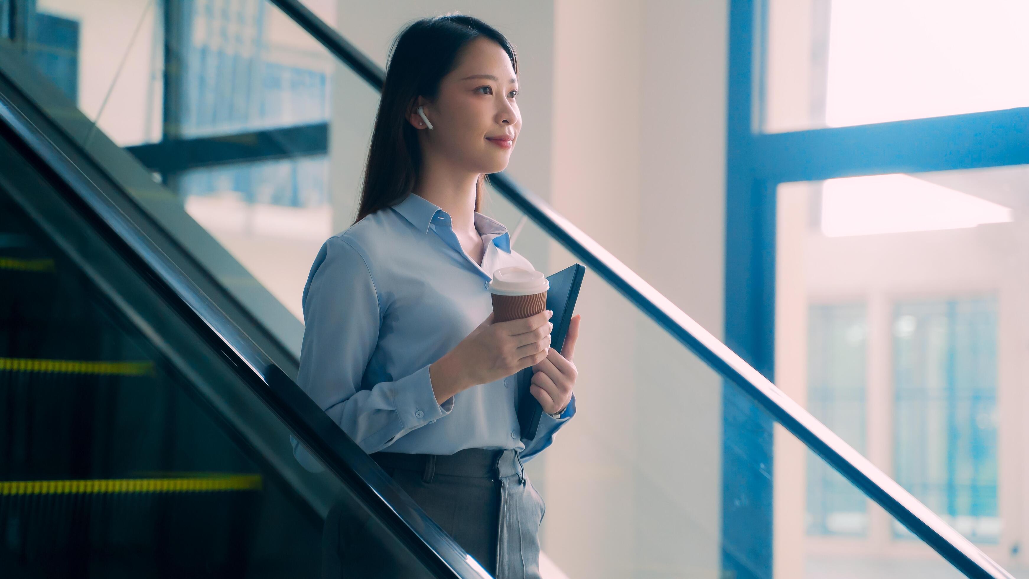 A business woman is walking down to escalator with a coffee cup in her hand Stock Free