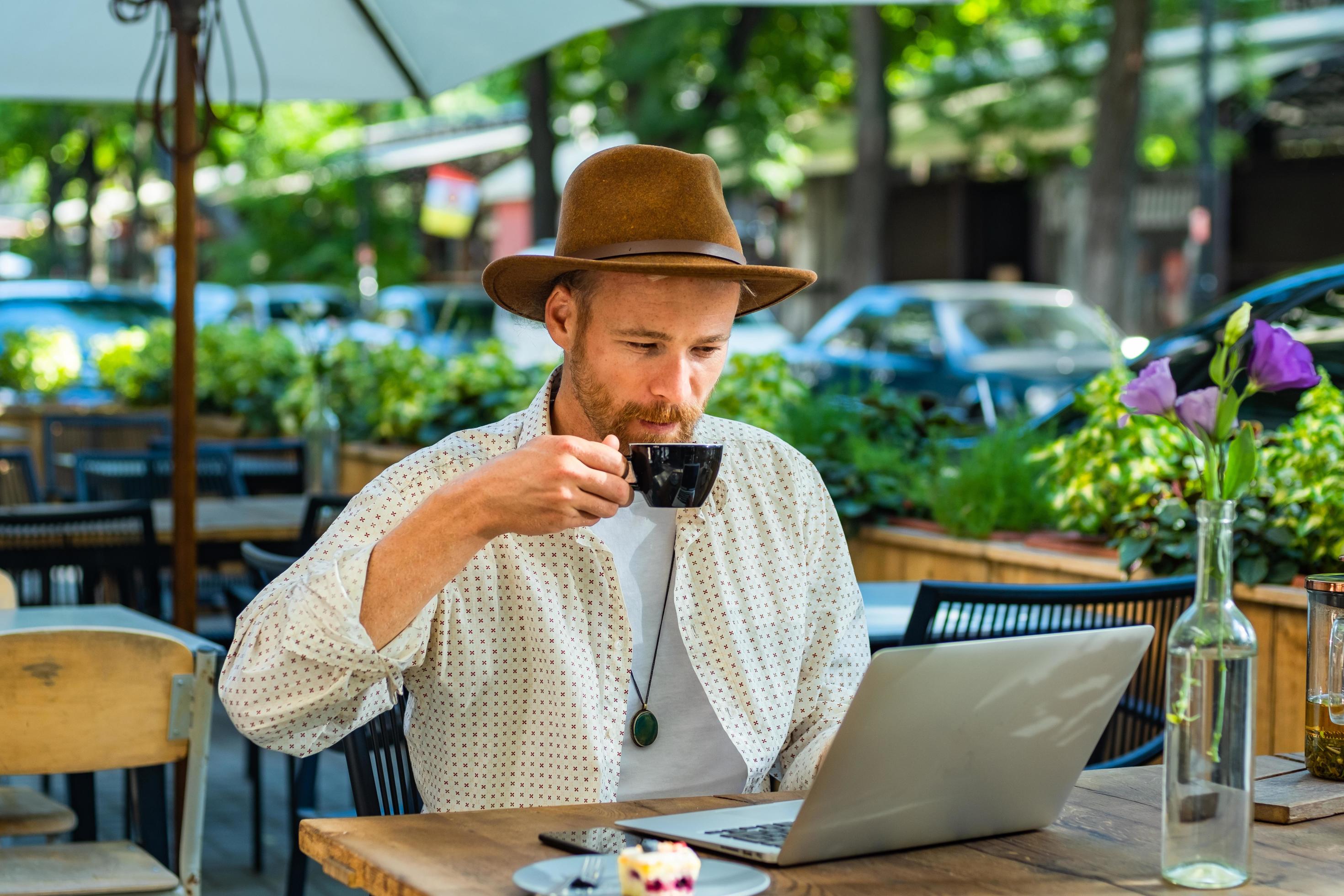 Young stylish hipster in hat work with laptop outdoors in the street cafe Stock Free