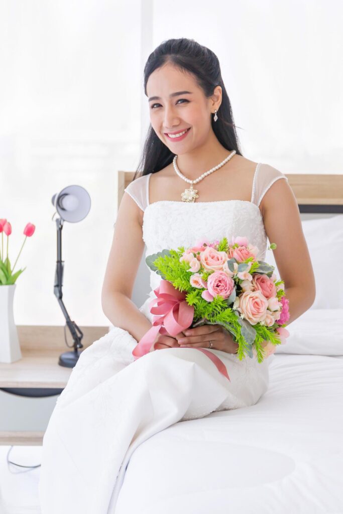 An Asian bride in a wedding dress is sitting smiling brightly on the bed in her hand holding a beautiful bouquet of flowers. Stock Free