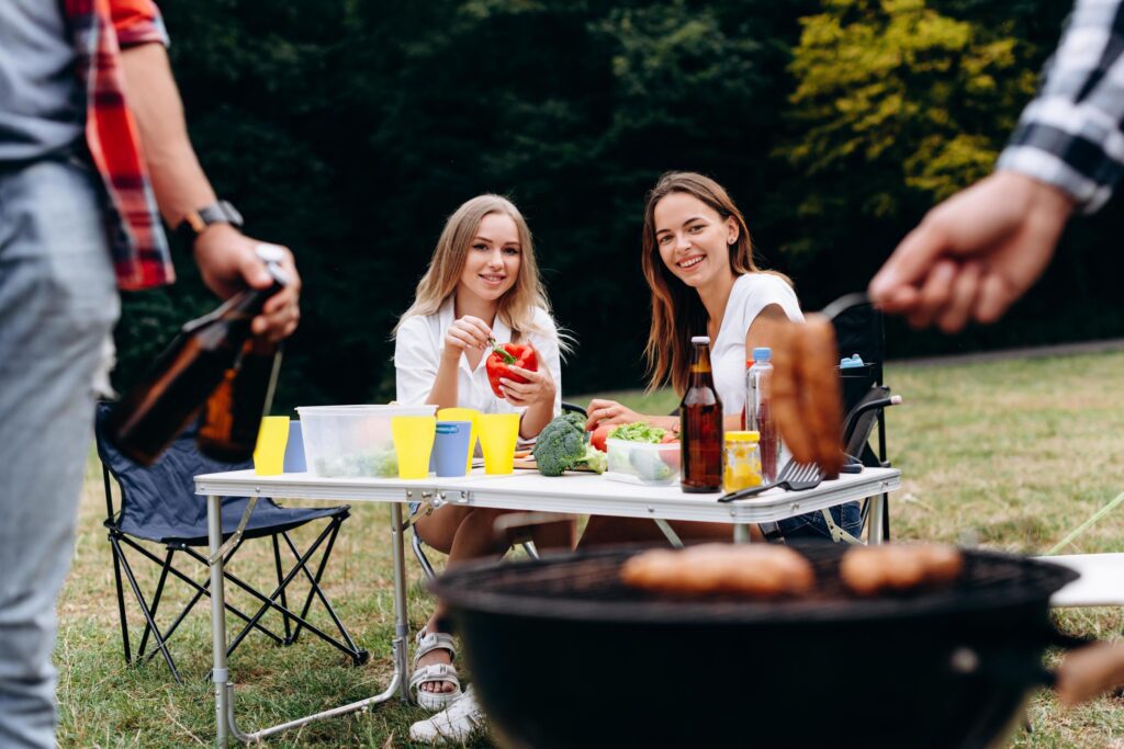 Smiling women sitting at the table with fresh food and looking on the barbecue Stock Free