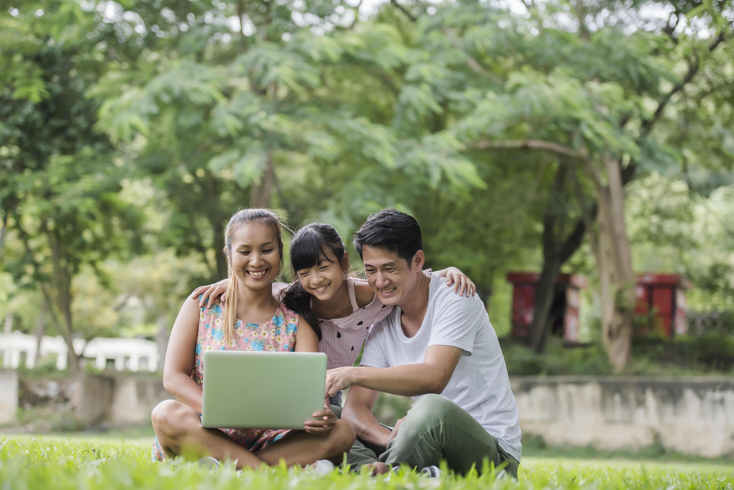 Happy family father, mother and daughter sitting on the grass and playing laptop at outdoor park Stock Free