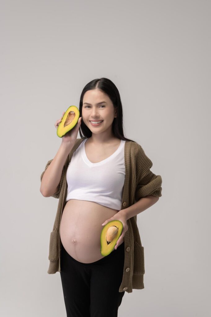 Portrait of Beautiful pregnant woman holding avocado over white background studio, health and maternity concept Stock Free