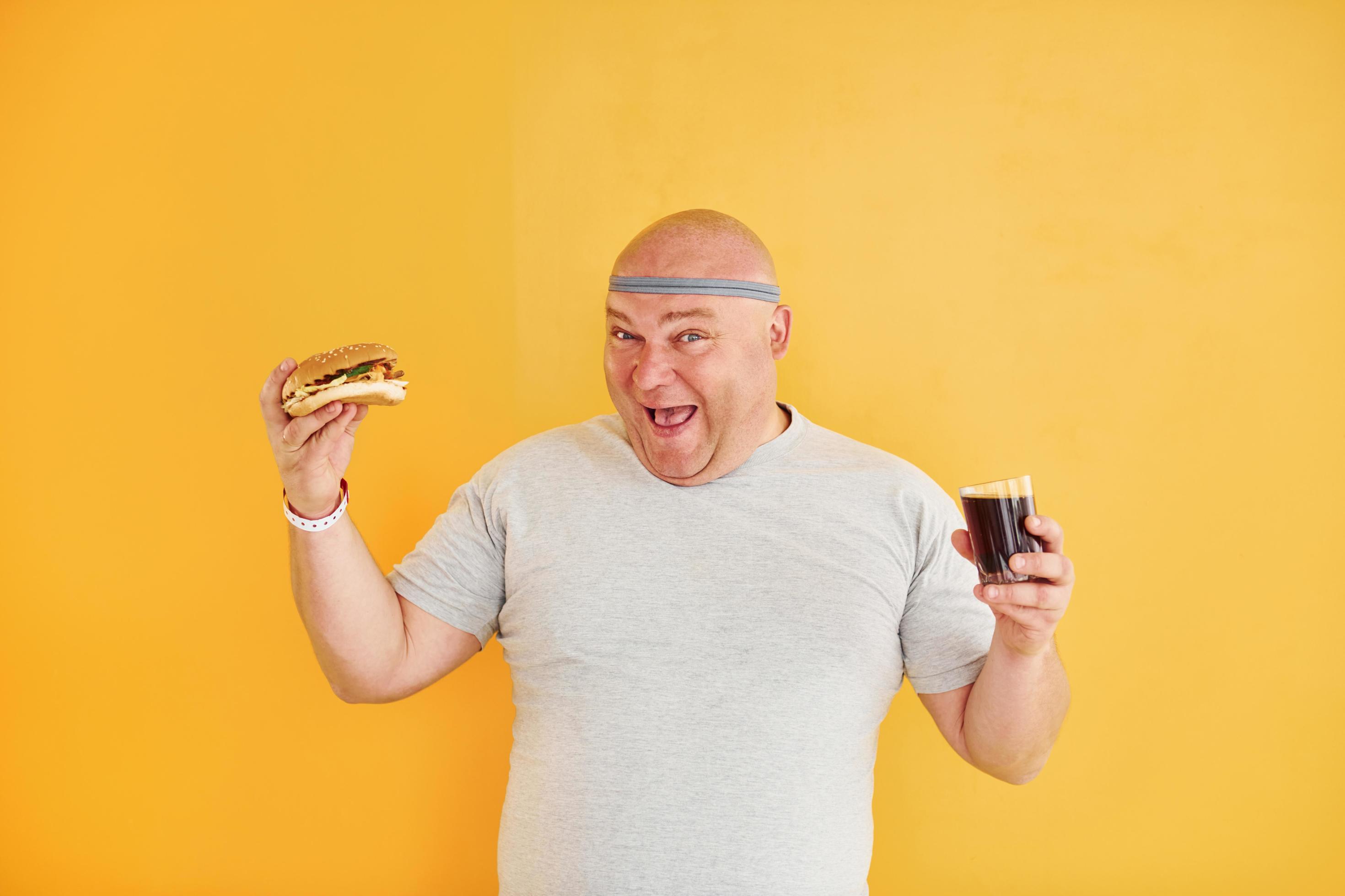 With fast food. Funny overweight man in sportive head tie is against yellow background Stock Free