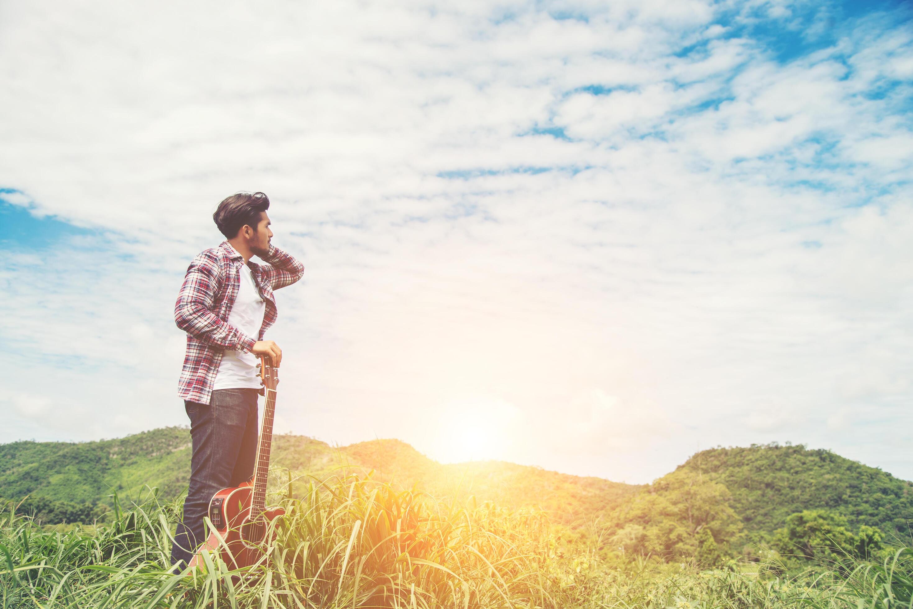 Young hipster man holding a guitar with a walking in nature, Relaxing in the field in a sunny blue sky day. Stock Free