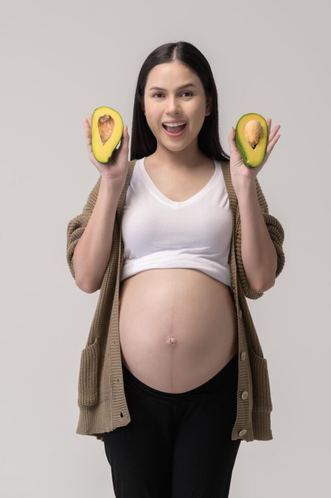 Portrait of Beautiful pregnant woman holding avocado over white background studio, health and maternity concept Stock Free