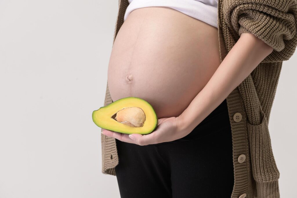 Portrait of Beautiful pregnant woman holding avocado over white background studio, health and maternity concept Stock Free