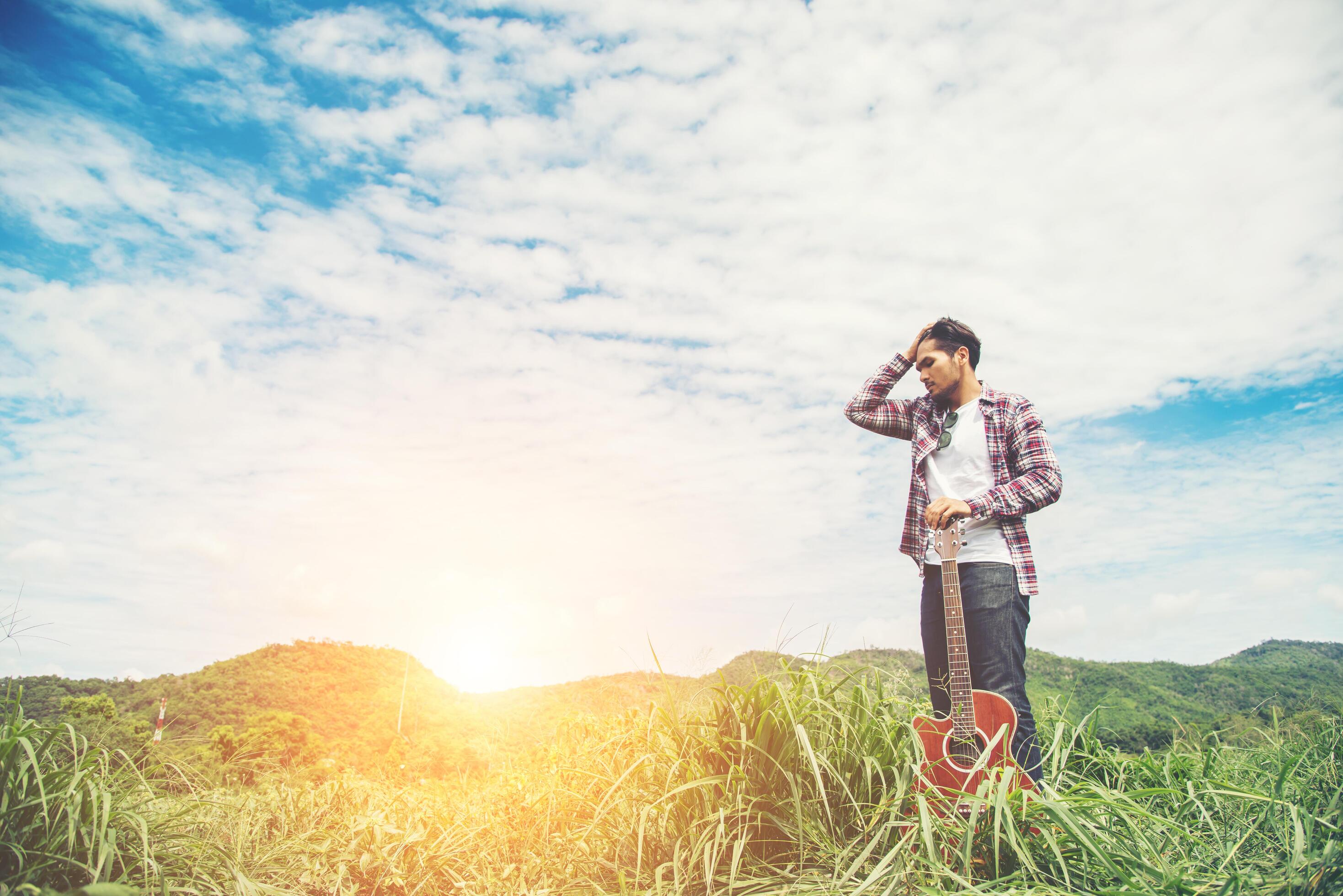 Young hipster man holding a guitar with a walking in nature, Relaxing in the field in a sunny blue sky day. Stock Free