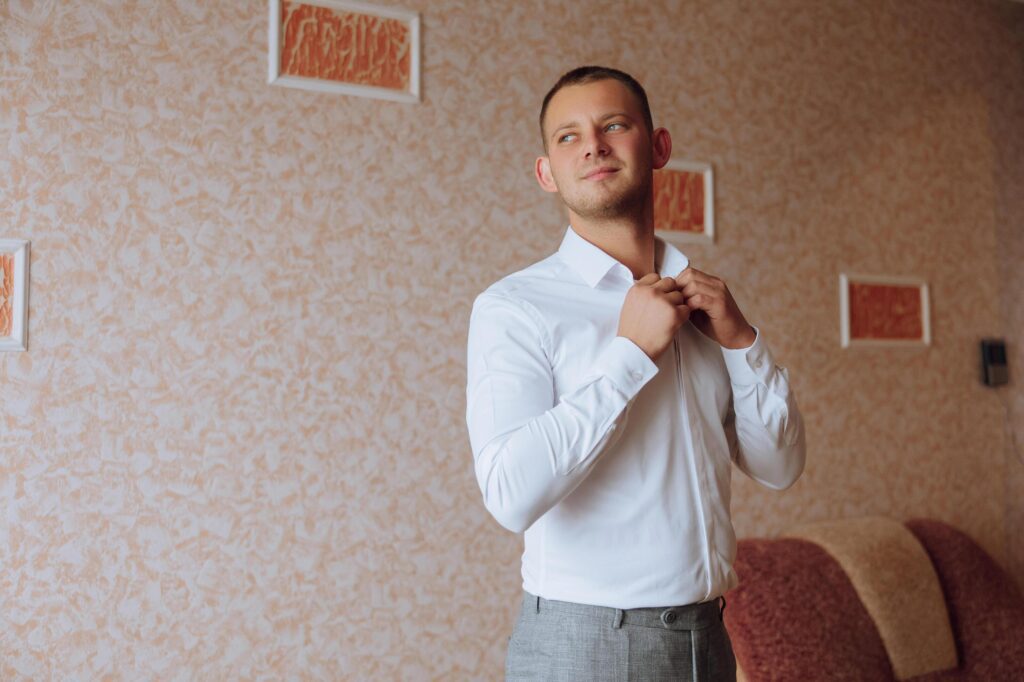 A man in a white shirt stands by the window in the room and fastens the buttons on his collar and sleeves. Watch on hand. Stylish business portrait of a man, close-up photo. The groom is preparing. Stock Free