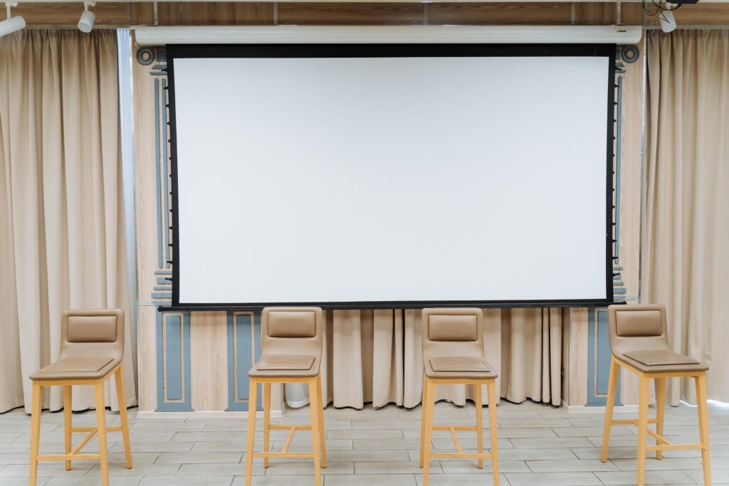 A white screen for showing the video hangs on the wall. Four chairs stand on the stage. Conference hall, business class for performances. Stock Free