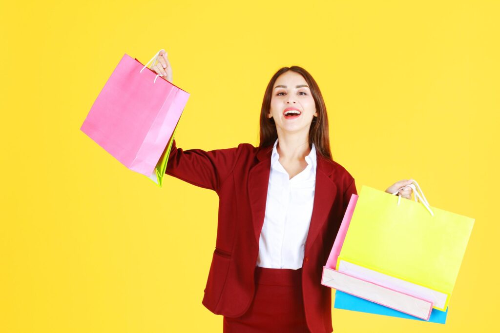 Beautiful working woman in red suit smile, happy and hand holding colorful shopping paper bags isolated on yellow background. Stock Free