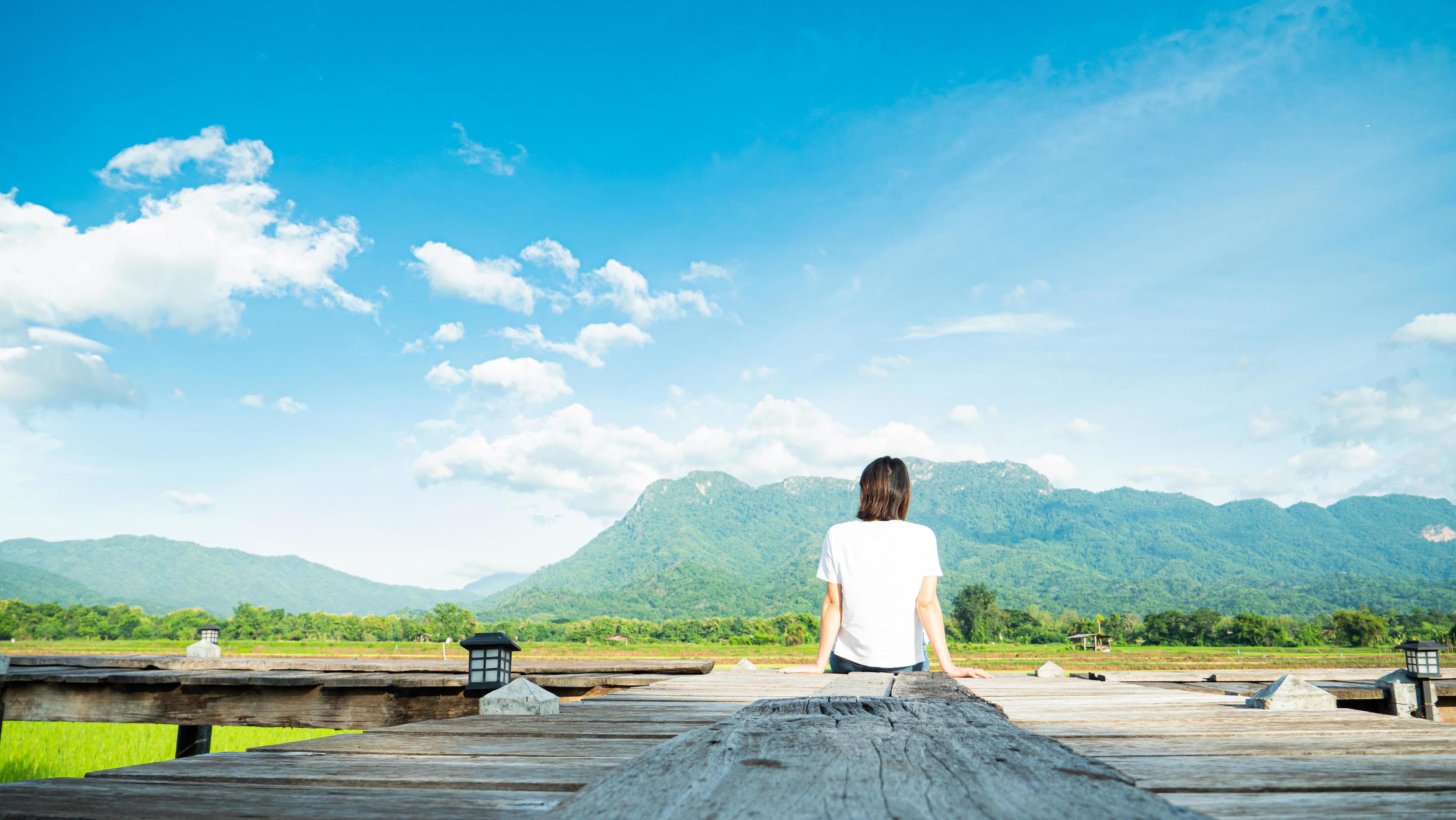 girl wearing a casual t-shirt Sit on a wooden walkway, see nature, mountains, rice fields, fresh air. and the morning sun bright blue sky white clouds Stock Free