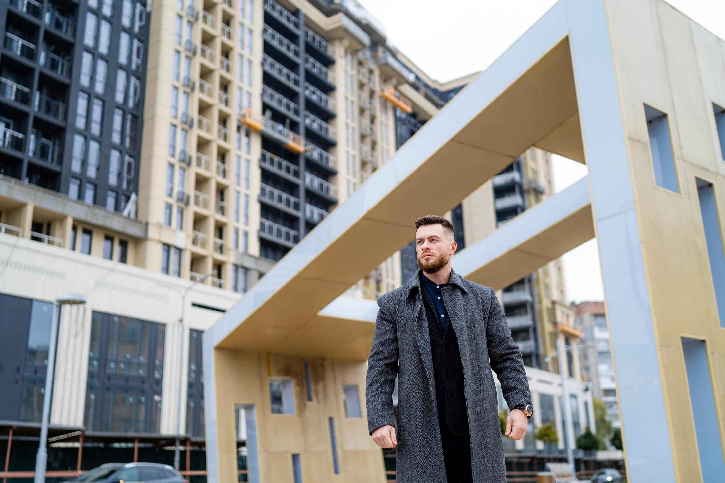 Trendy handsome man in gray coat and dark suit poses on modern architecture background. Beautiful photo zone. Stock Free