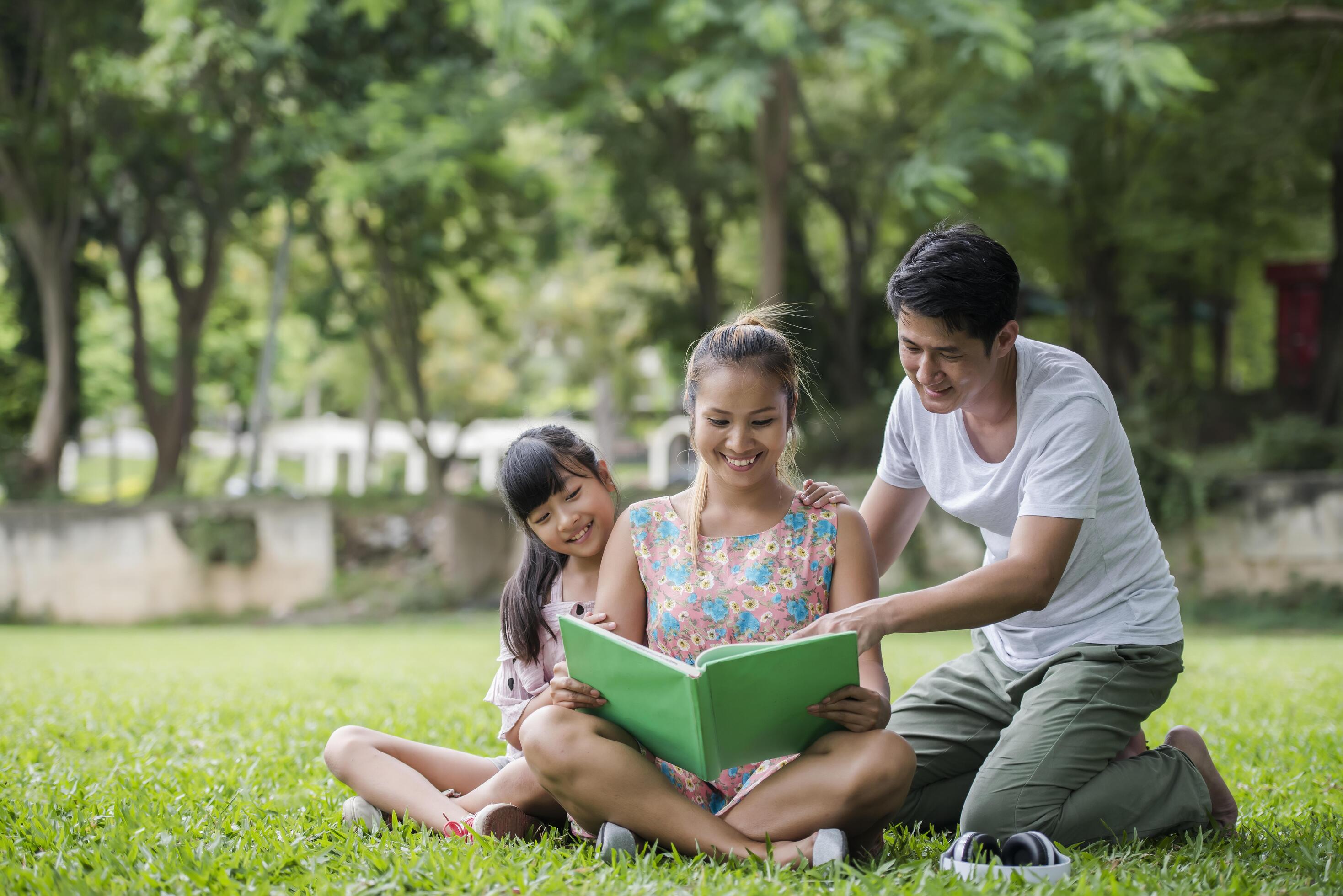 Happy Family mother, father and daughter read a book in the park Stock Free