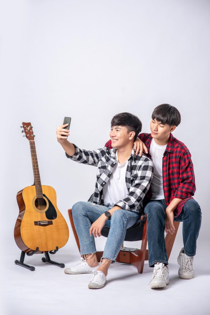 Two loving young men sit on a chair and take a selfie from a smartphone. Free Photo