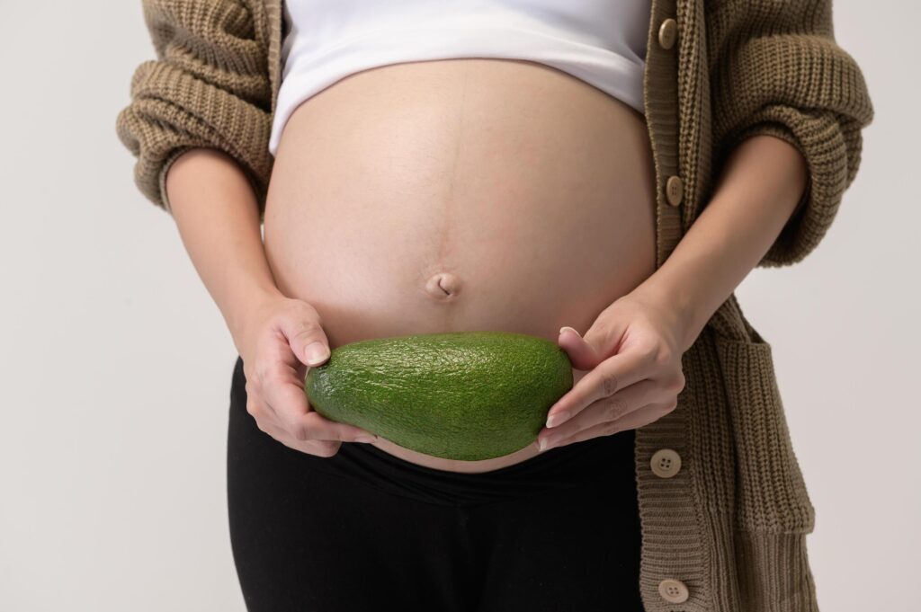 Portrait of Beautiful pregnant woman holding avocado over white background studio, health and maternity concept Stock Free