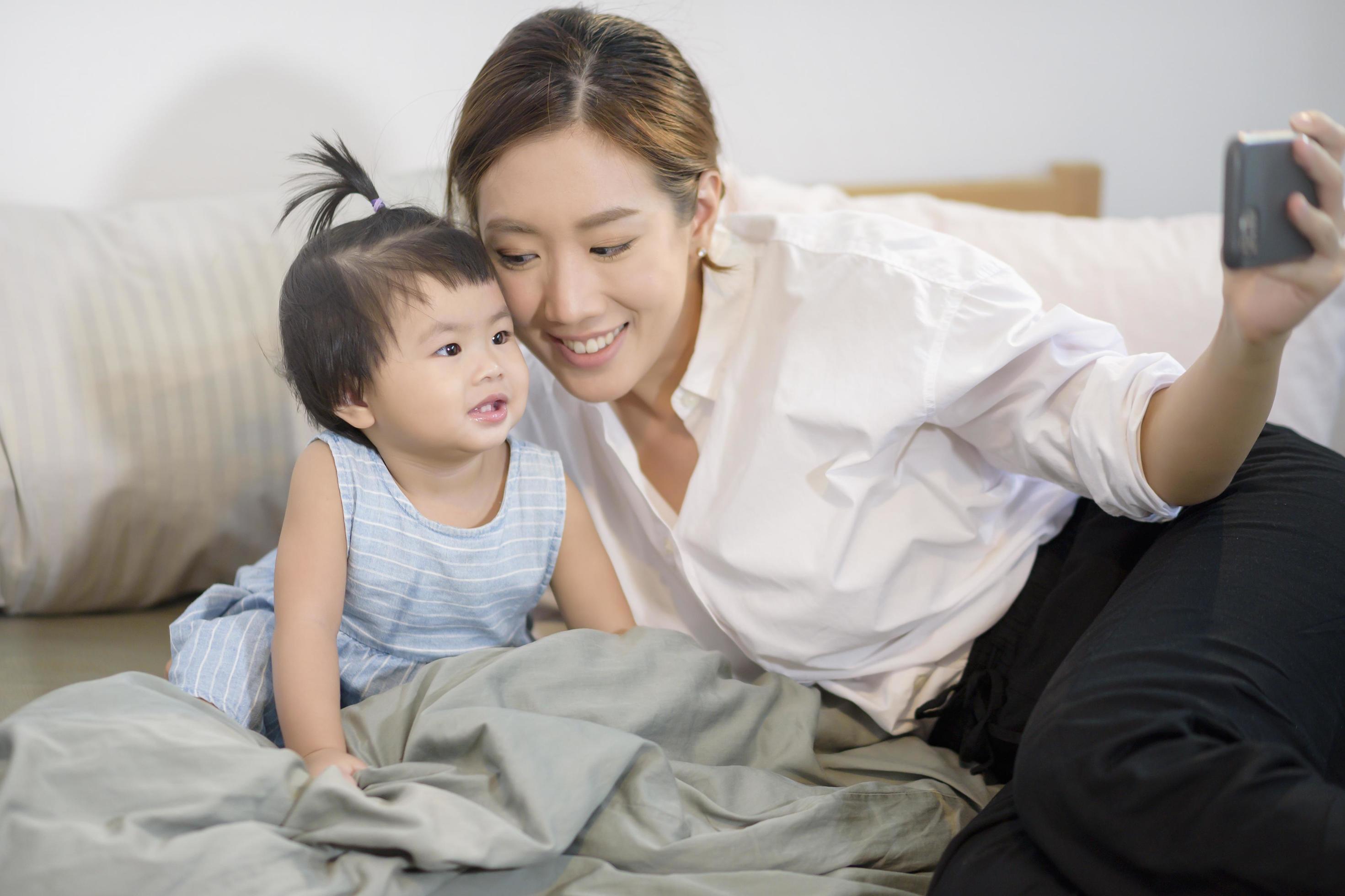 Asian Mother and her baby daughter are making selfie or video call to father in bed , Family , Home safety , parenthood , technology concept Stock Free