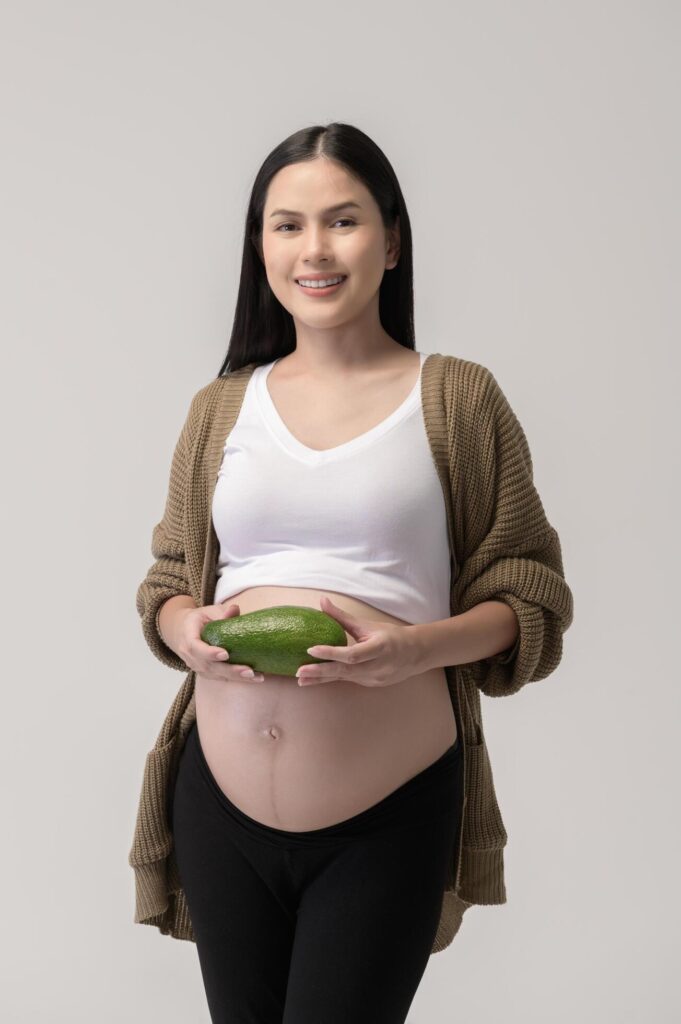 Portrait of Beautiful pregnant woman holding avocado over white background studio, health and maternity concept Stock Free