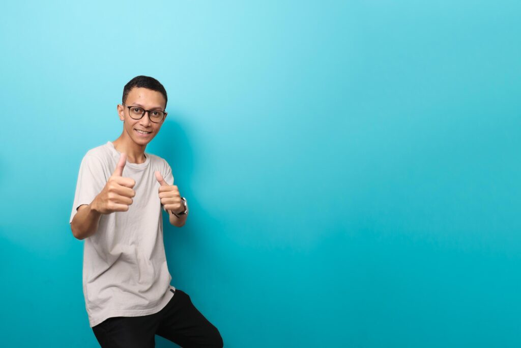 an Asian man in a plain white t-shirt with glasses with a like or best gesture isolated on blue background Stock Free