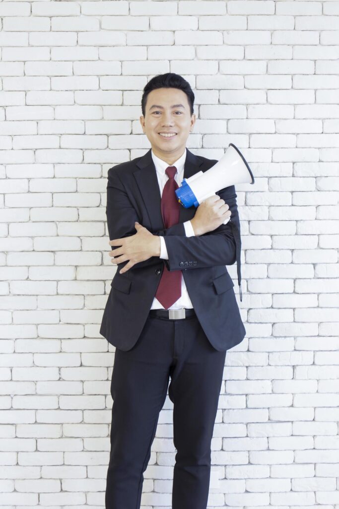 An Asian man in a suit holds a megaphone on a white brick wall. Stock Free