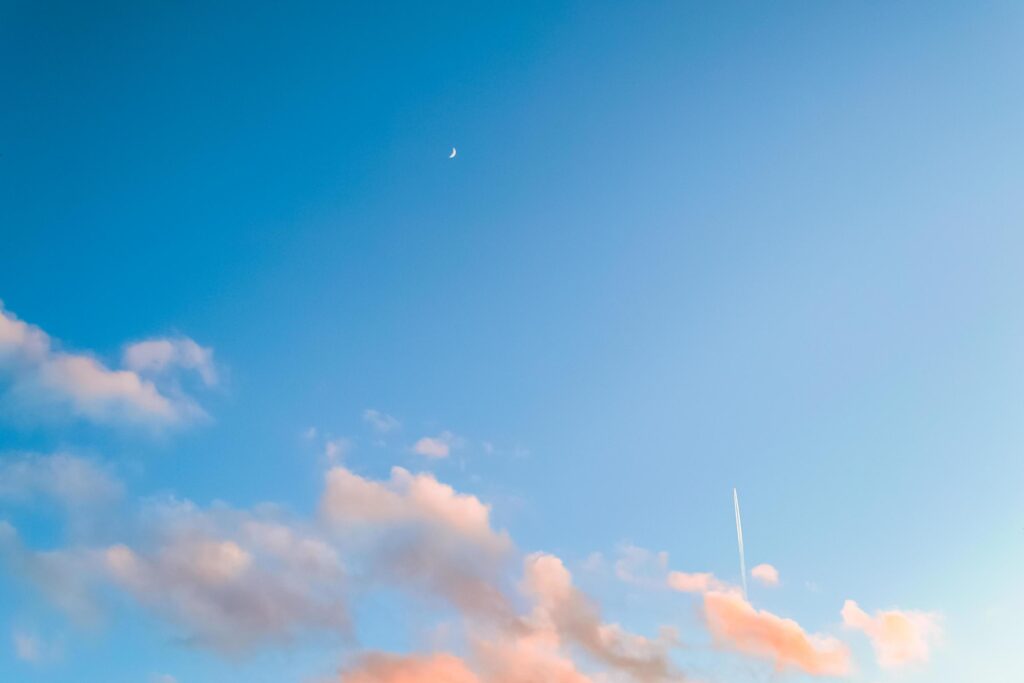 Nature background of blue sky with pink clouds, airplane trail, and early moon Stock Free