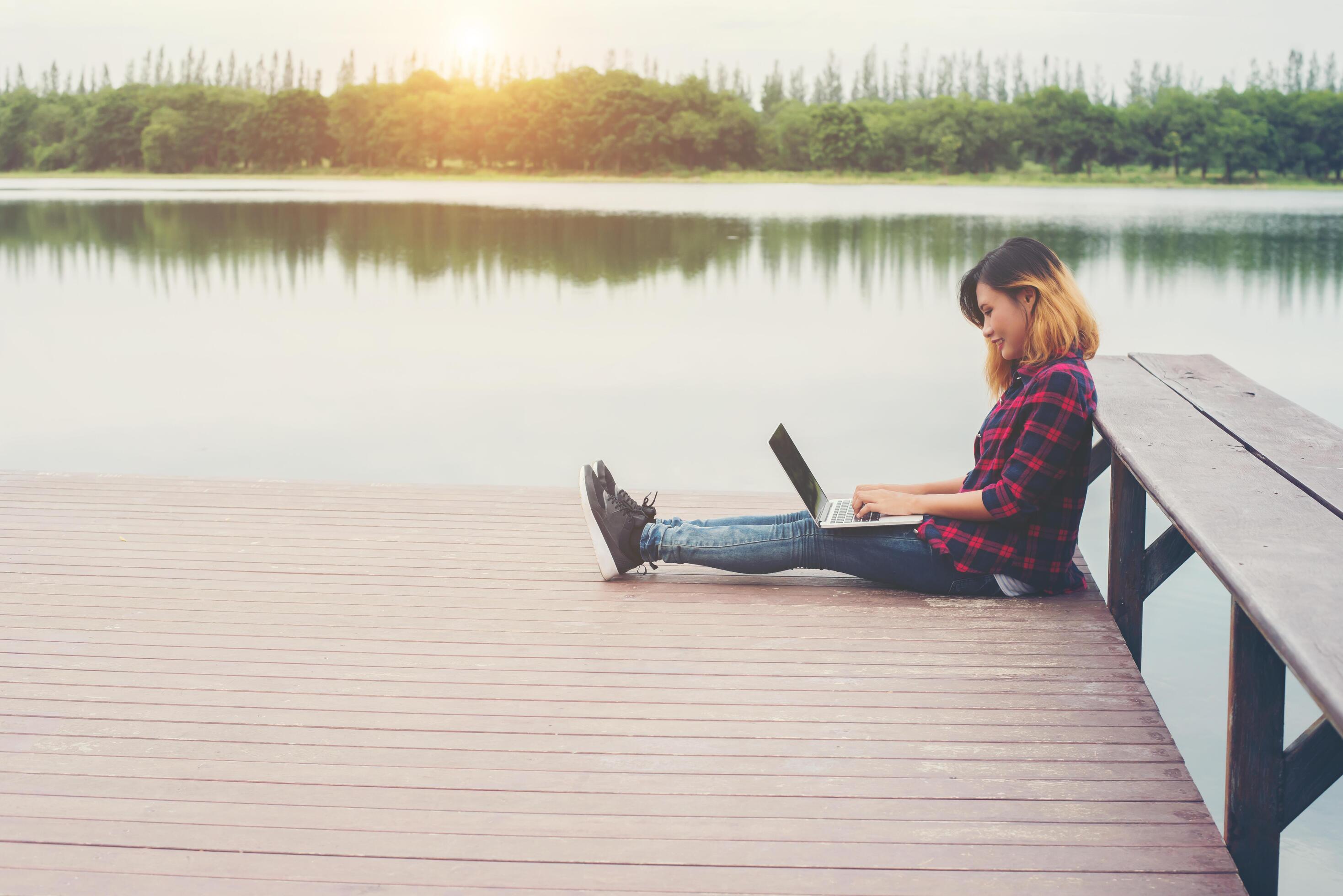 Young happy hipster woman working with her laptop sitting on pier,relaxing enjoy with nature. Stock Free