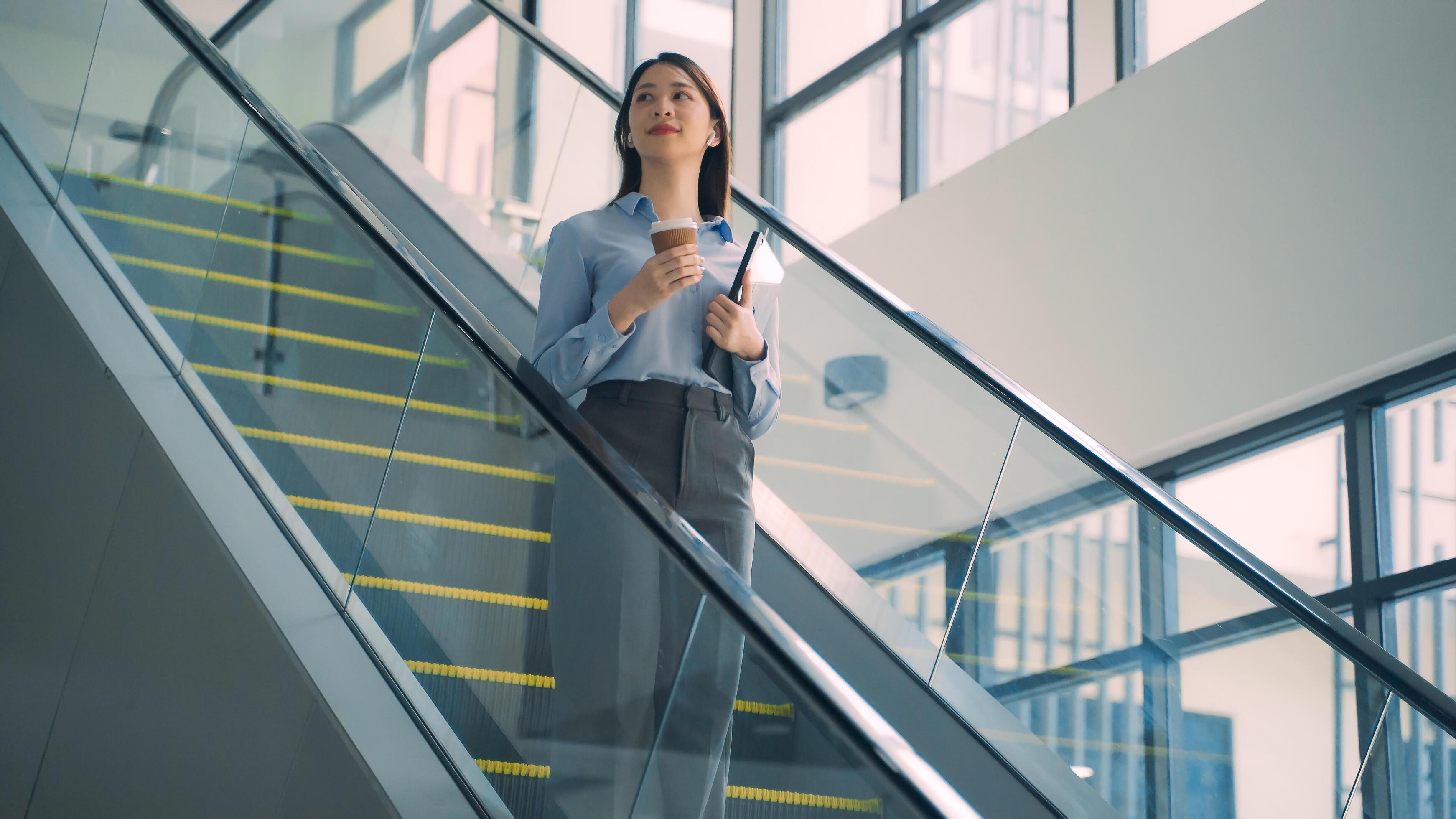 A business woman is walking down to escalator with a coffee cup in her hand Stock Free