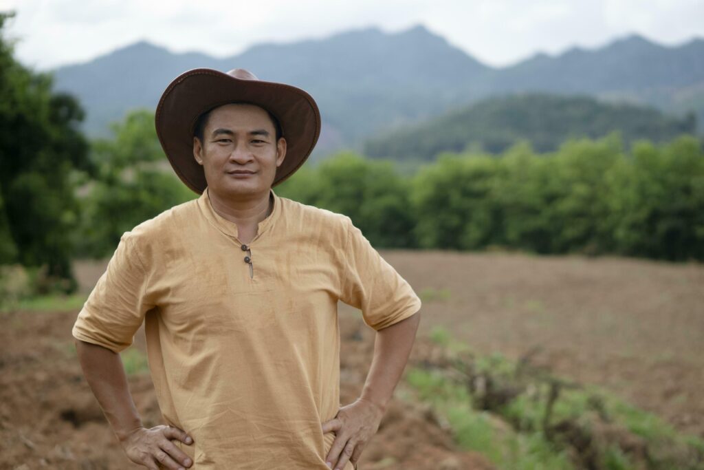 Handsome Asian man farmer wears hat, brown shirt, put hands on waist, feels confident, stands at agriculture land before growing crops. Concept, agriculture occupation. Thai farmer lifestyle. Stock Free