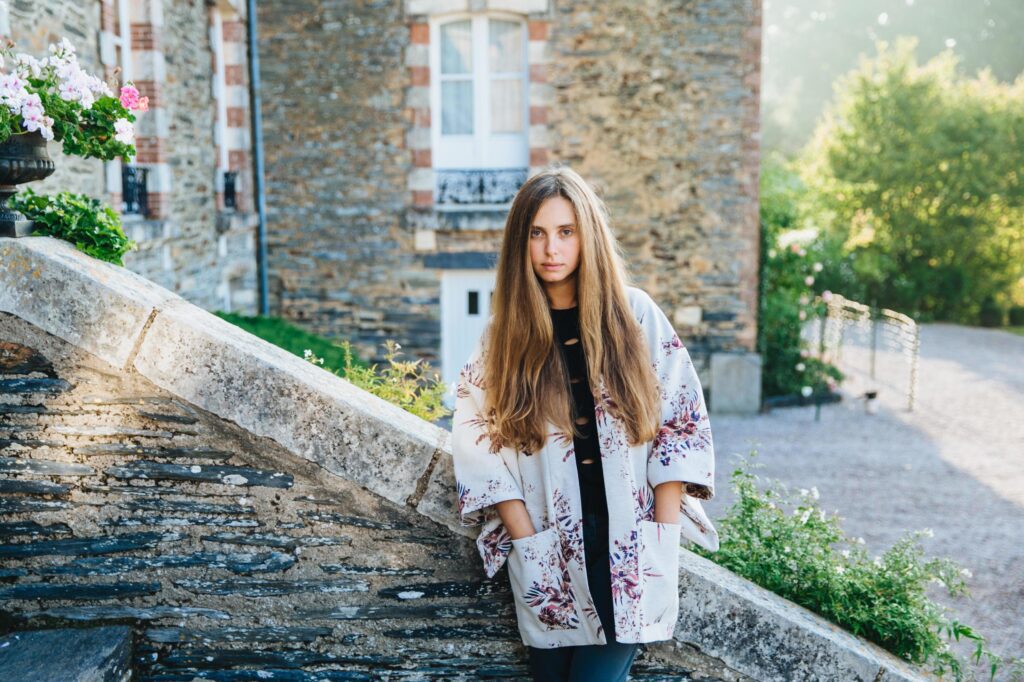 Outdoor portrait of attractive woman with long luxurious hair, wears fashionable mantel, stands on ancient stairs of builduing, poses seriously at camera. People, lifestyle, tourism concept. Stock Free