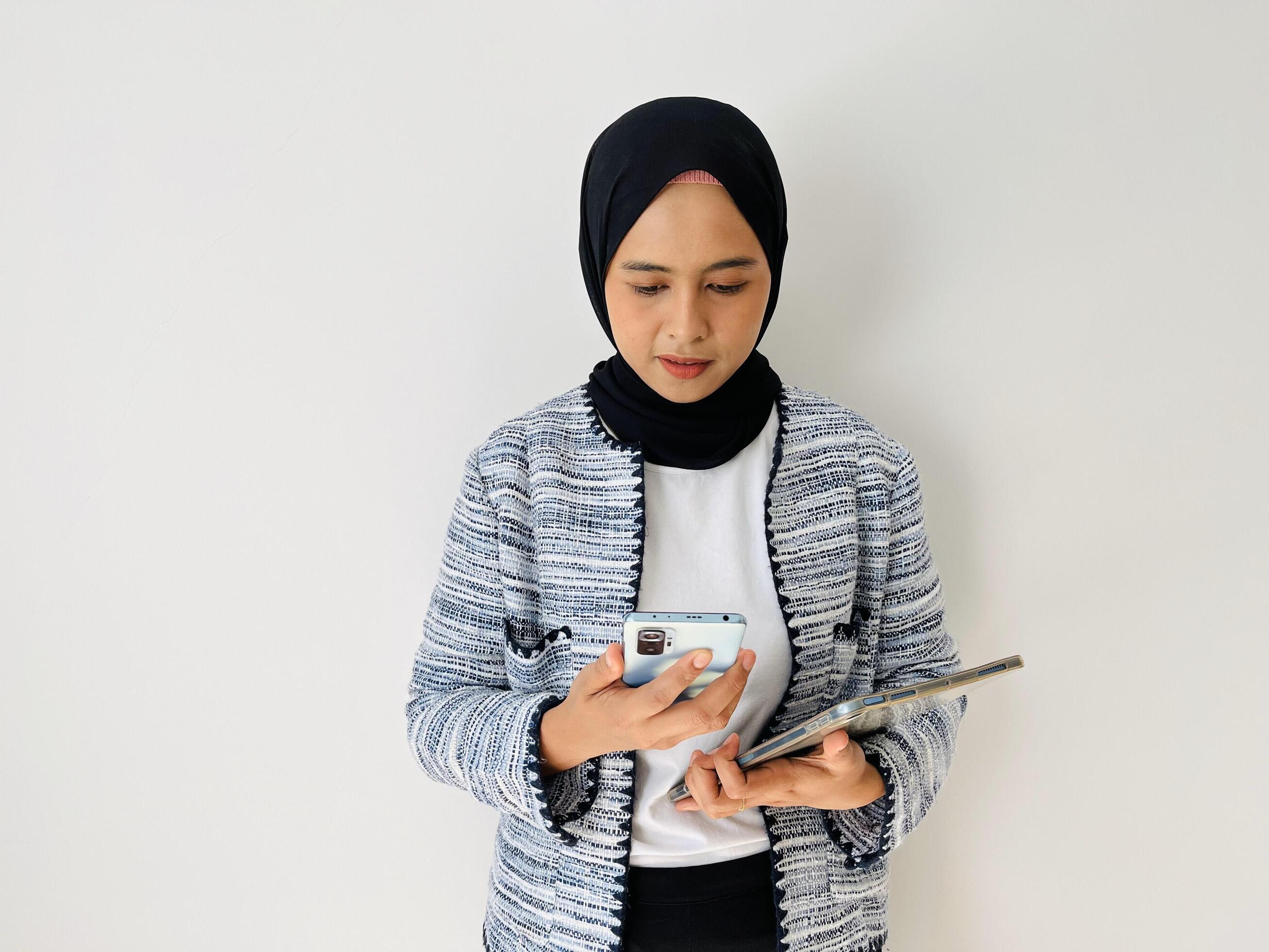 Young Asian woman smiling while using mobile phone. Standing female wearing black hijab and tweed blazer isolated on white background. Stock Free