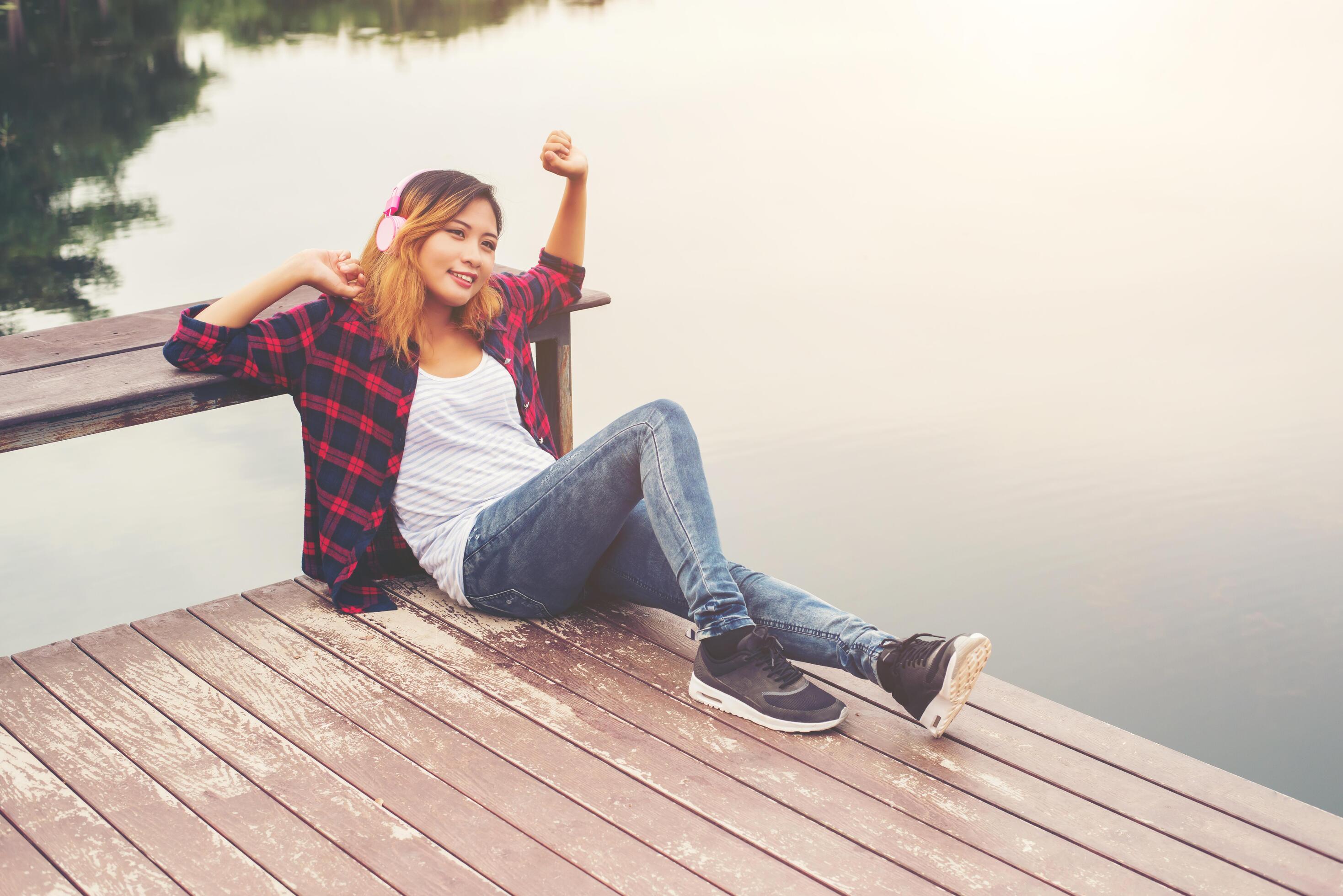 Portrait of young hipster woman with headphones sitting on pier and listening music relaxing with sunset,Lifestyle concept. Stock Free