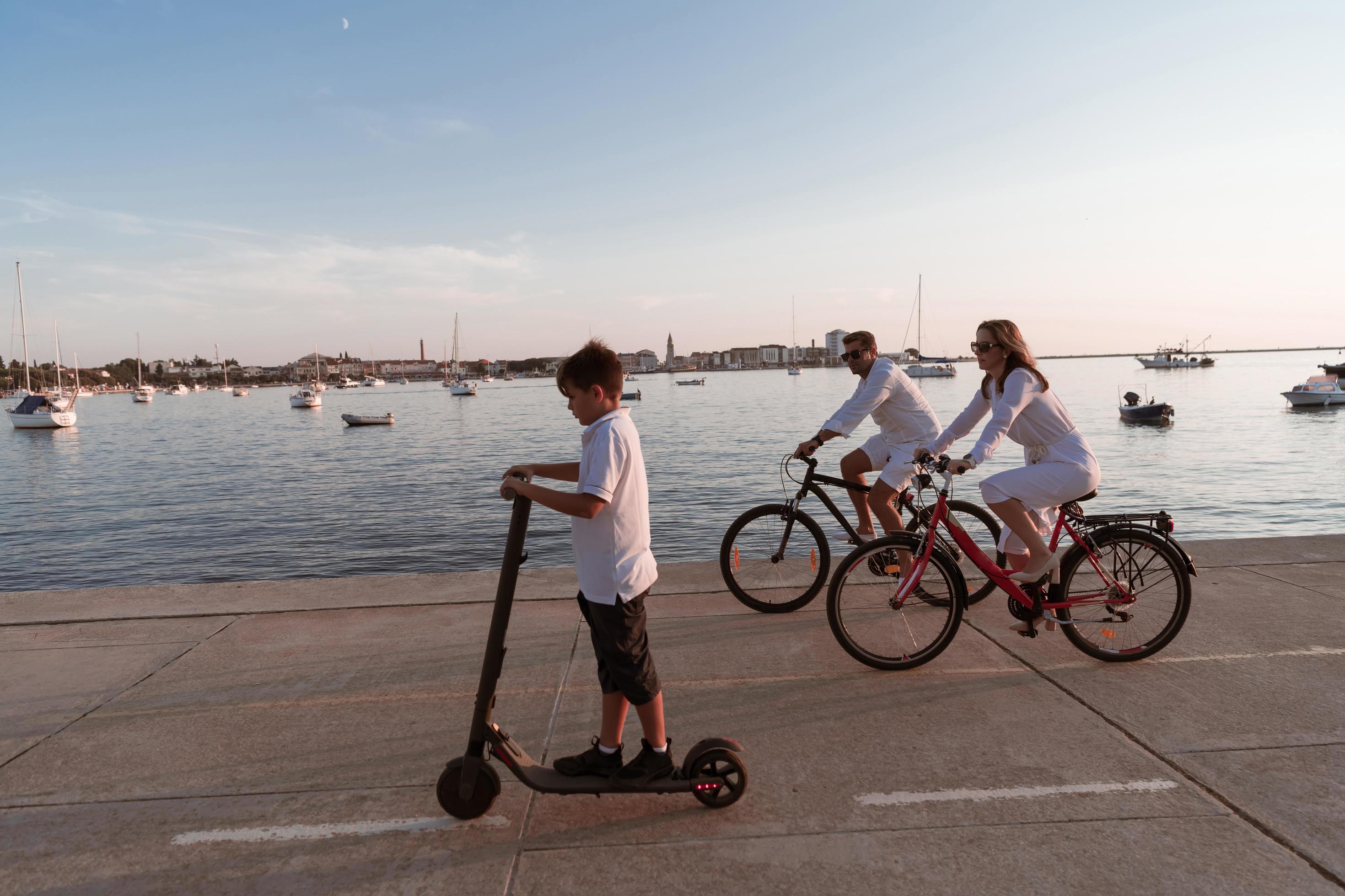 Happy family enjoying a beautiful morning by the sea together, parents riding a bike and their son riding an electric scooter. Selective focus Stock Free