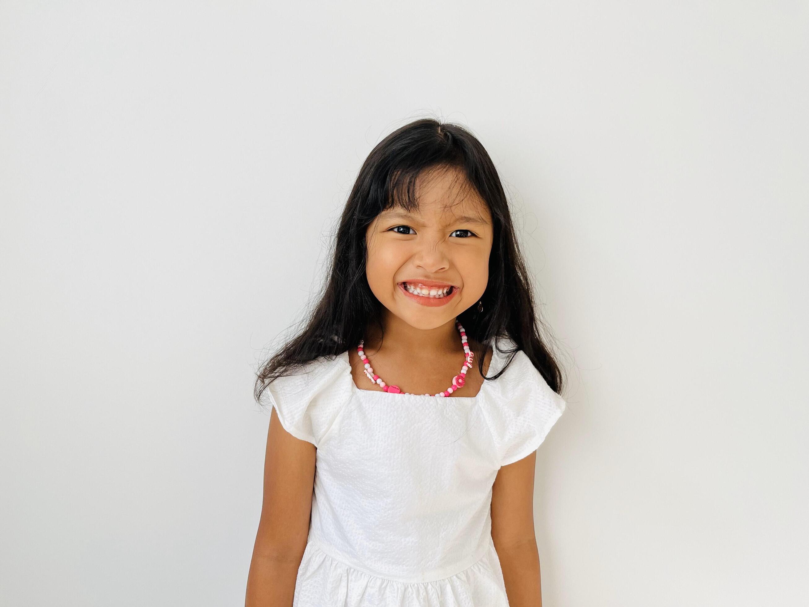 Portrait of a 5 year old Asian girl with shoulder length black hair wearing a white dress and necklace smiling broadly at the camera Stock Free