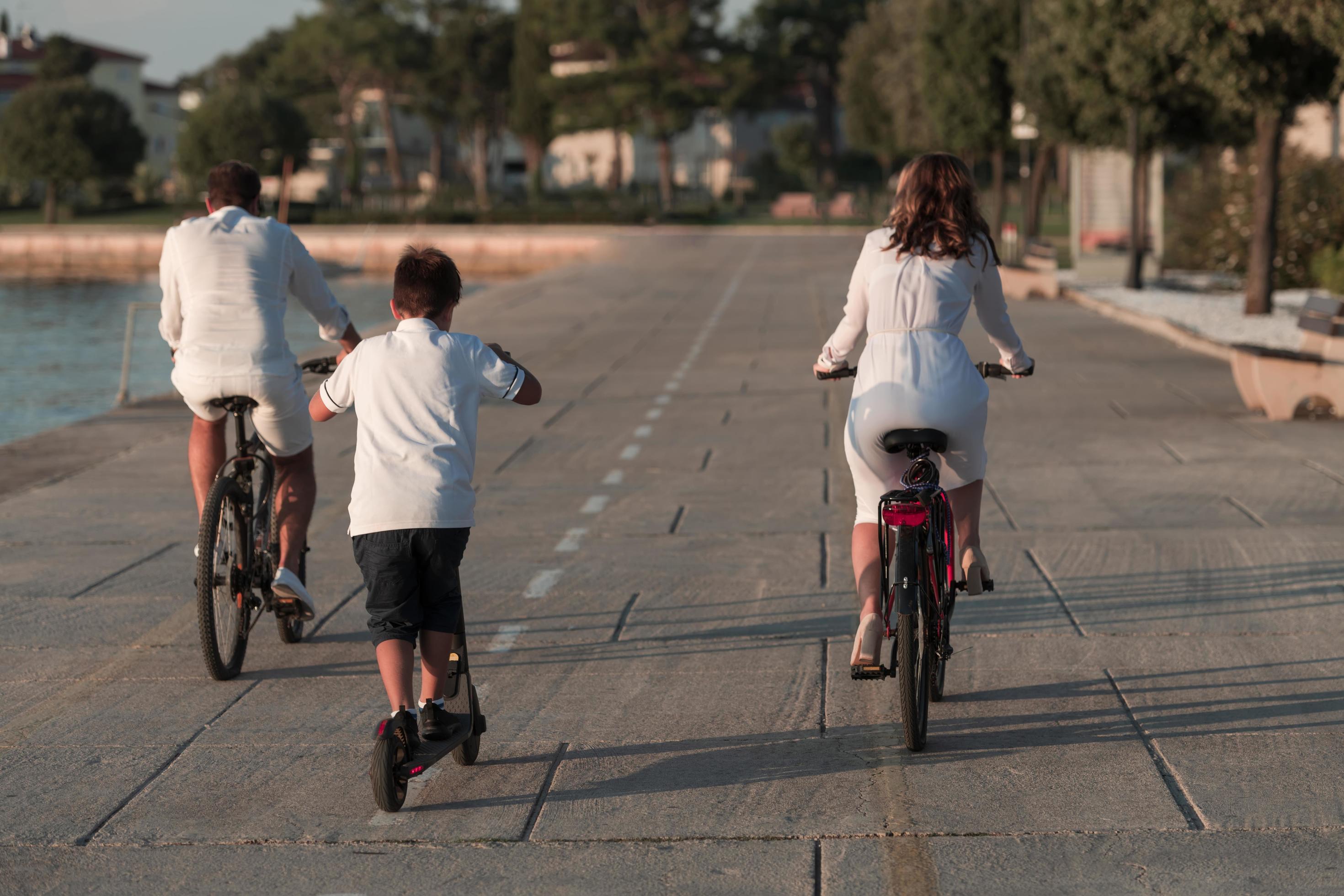 Happy family enjoying a beautiful morning by the sea together, parents riding a bike and their son riding an electric scooter. Selective focus Stock Free