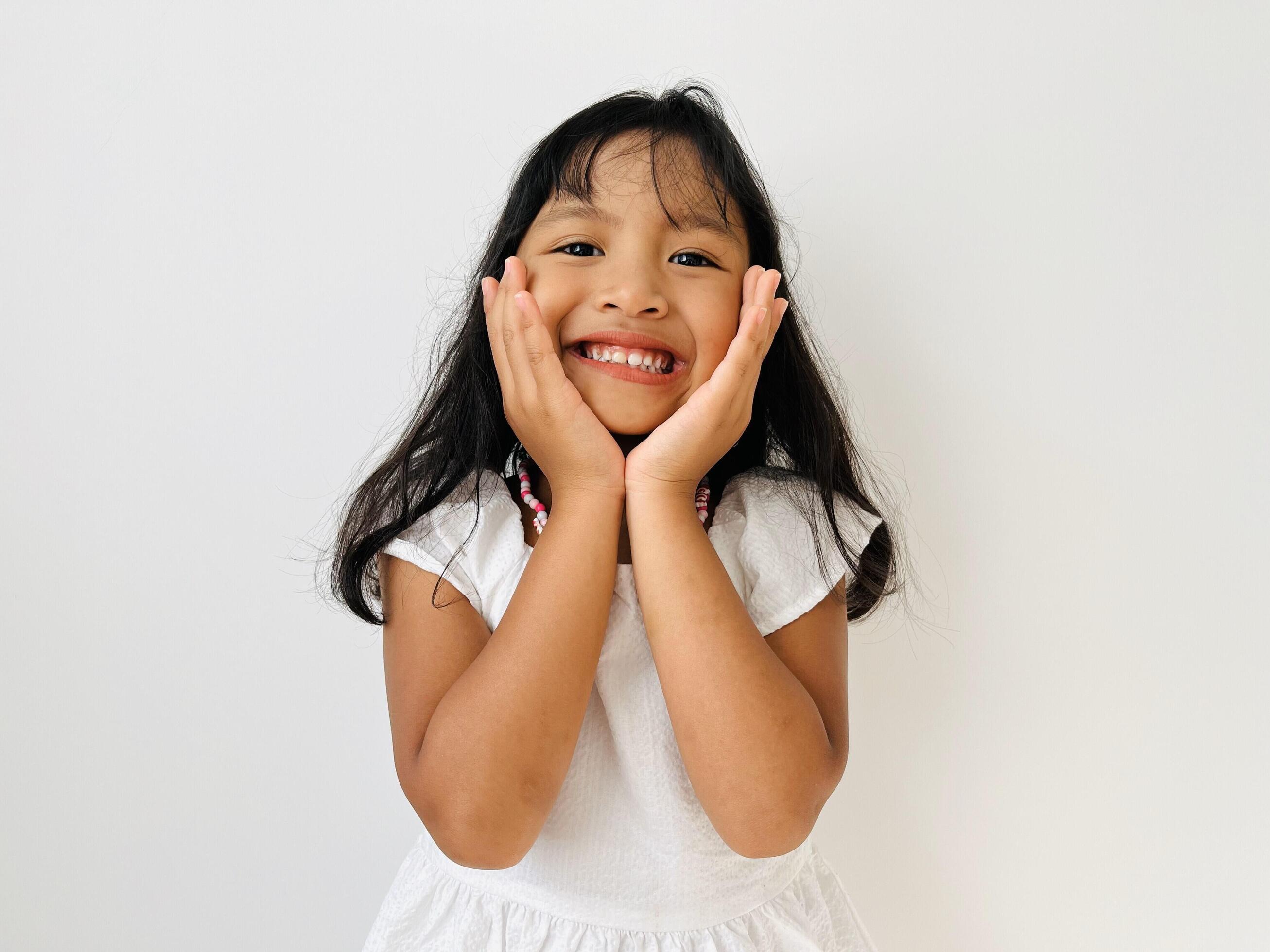 A cute 5 year old little girl wearing a white dress smiles broadly with both hands holding her chin Stock Free