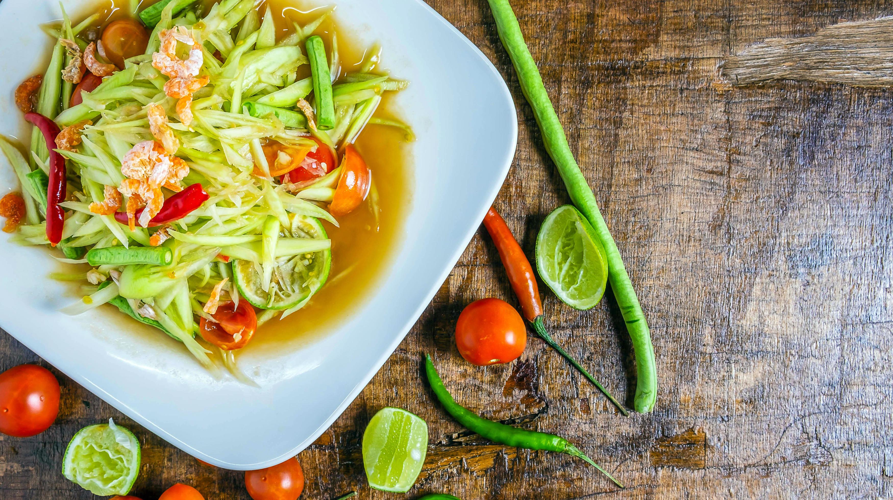 Thai food papaya salad on a plate with lemon and tomato on a wooden background.Top view Stock Free