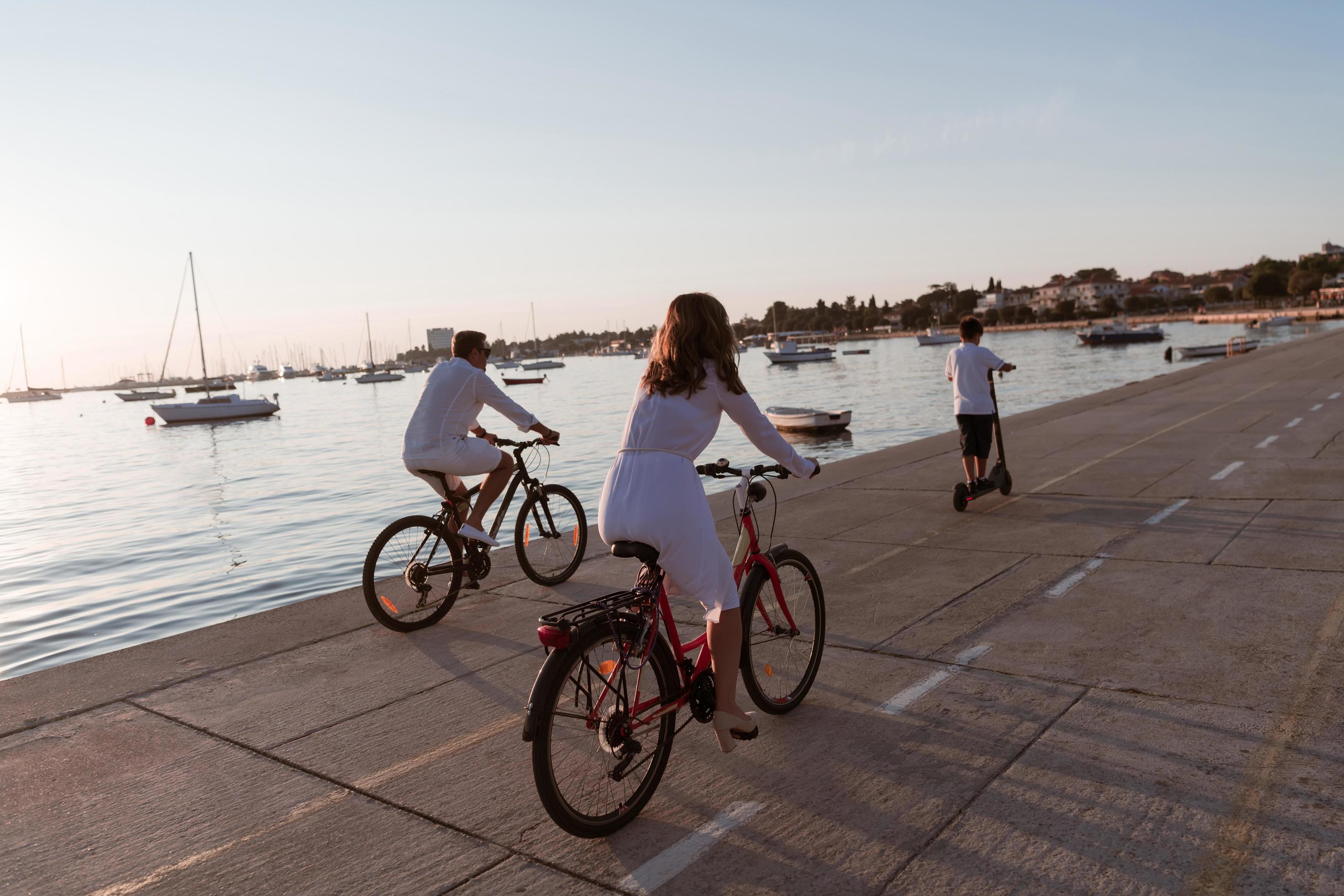 Happy family enjoying a beautiful morning by the sea together, parents riding a bike and their son riding an electric scooter. Selective focus Stock Free