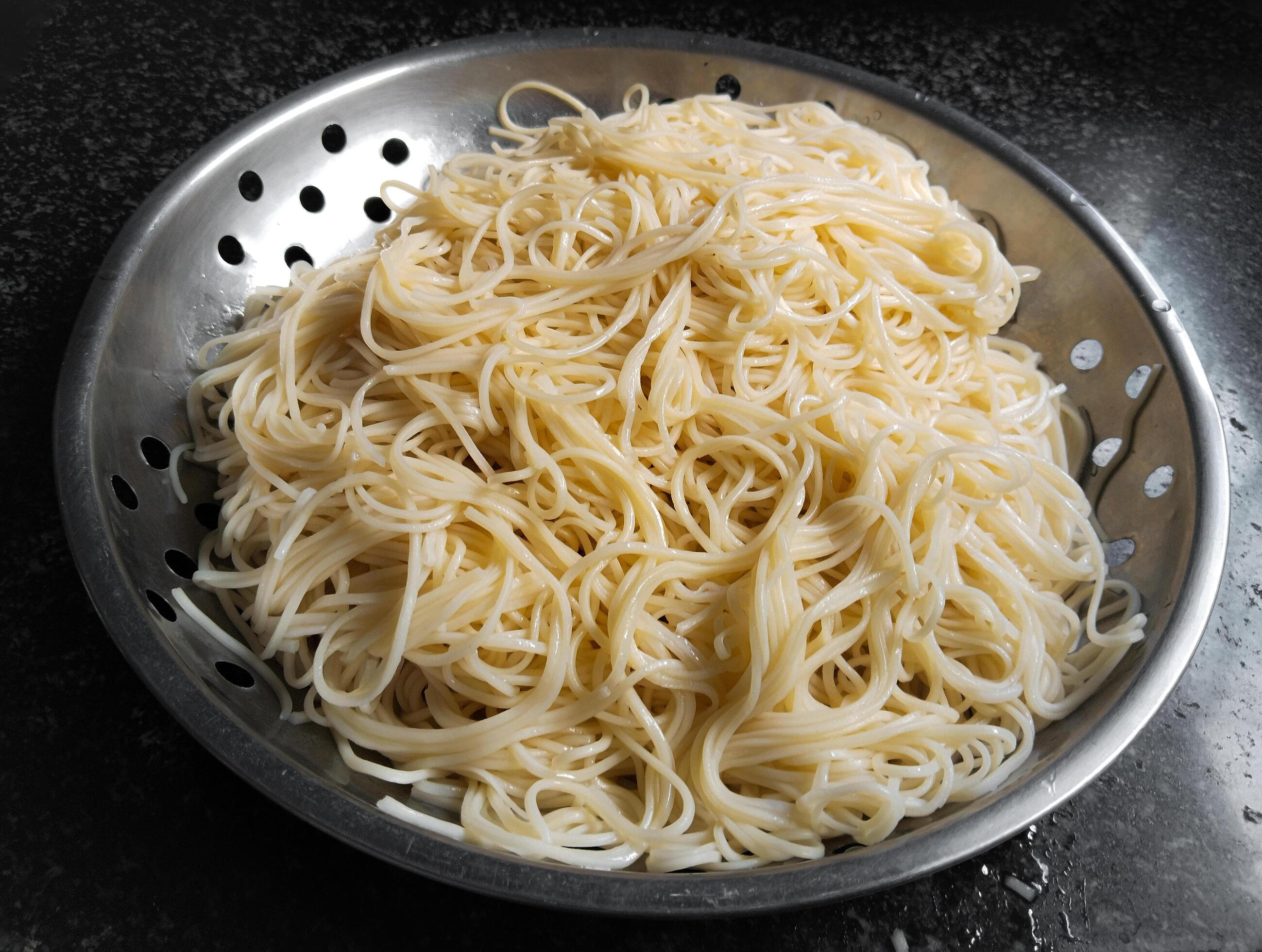 A colander filled with cooked egg noodles, placed on a dark counter top Stock Free