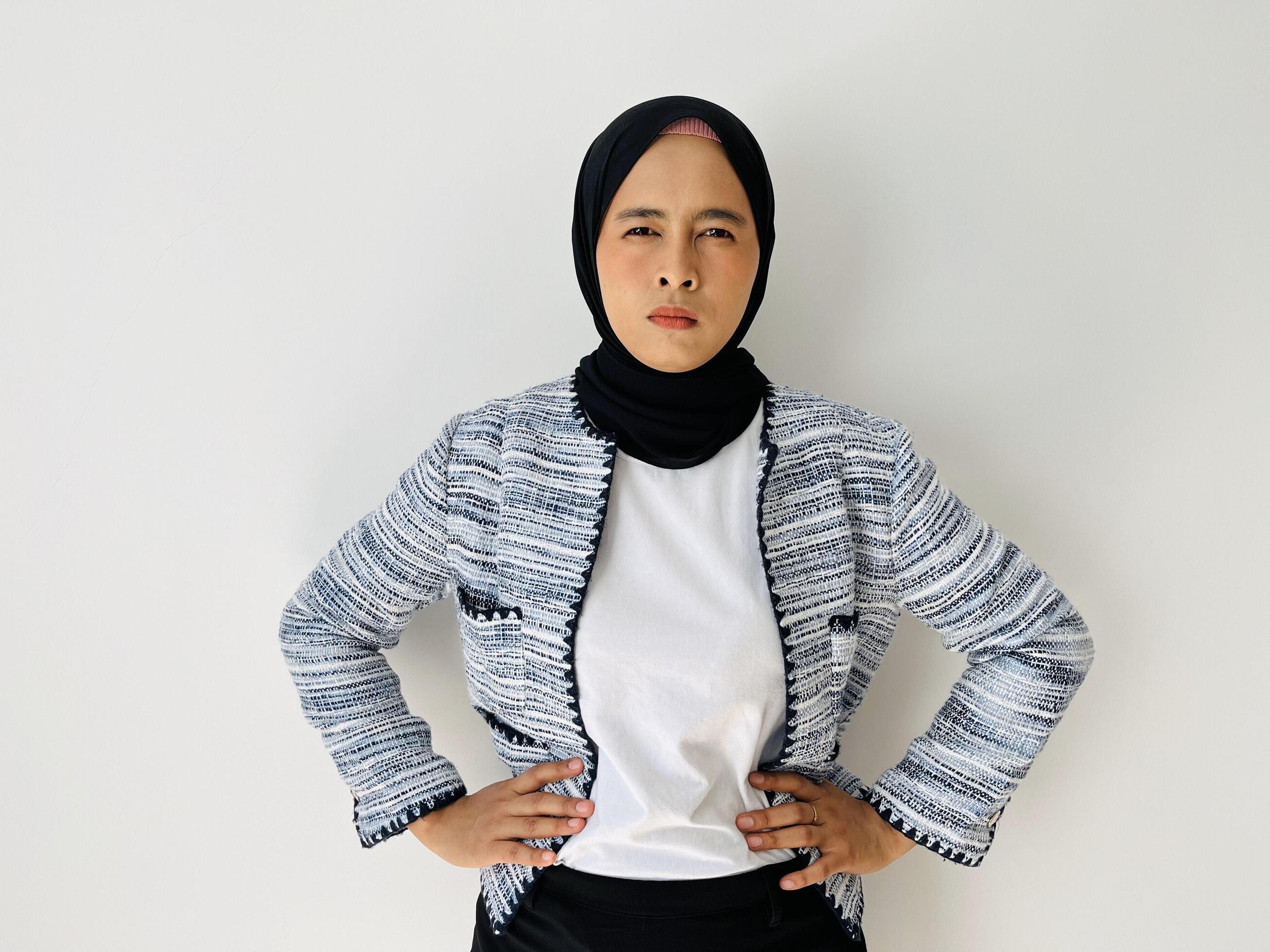 Angry young Asian woman with hand in her waist. Standing female wearing black hijab and tweed blazer isolated on white background. Stock Free