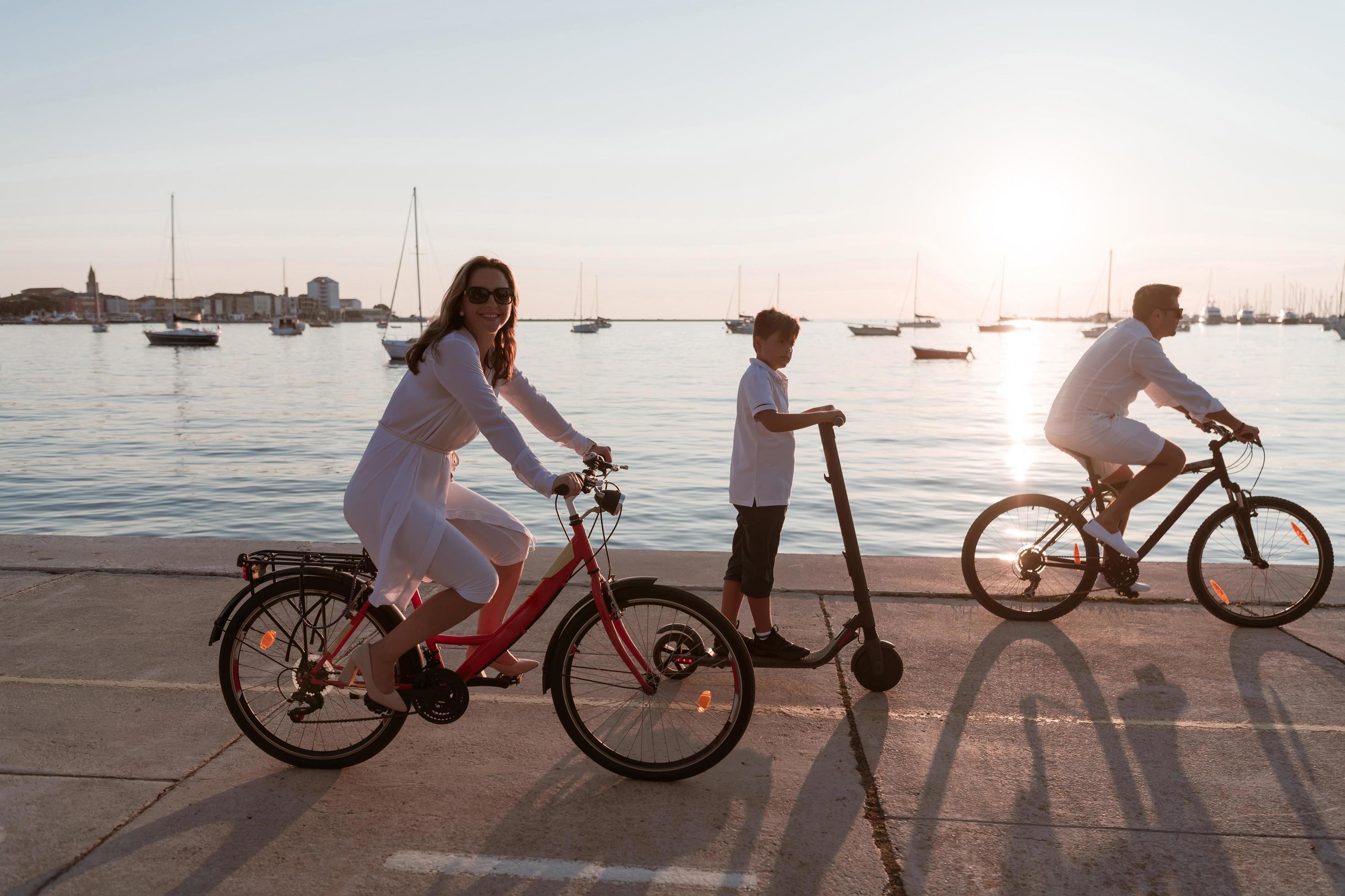 Happy family enjoying a beautiful morning by the sea together, parents riding a bike and their son riding an electric scooter. Selective focus Stock Free