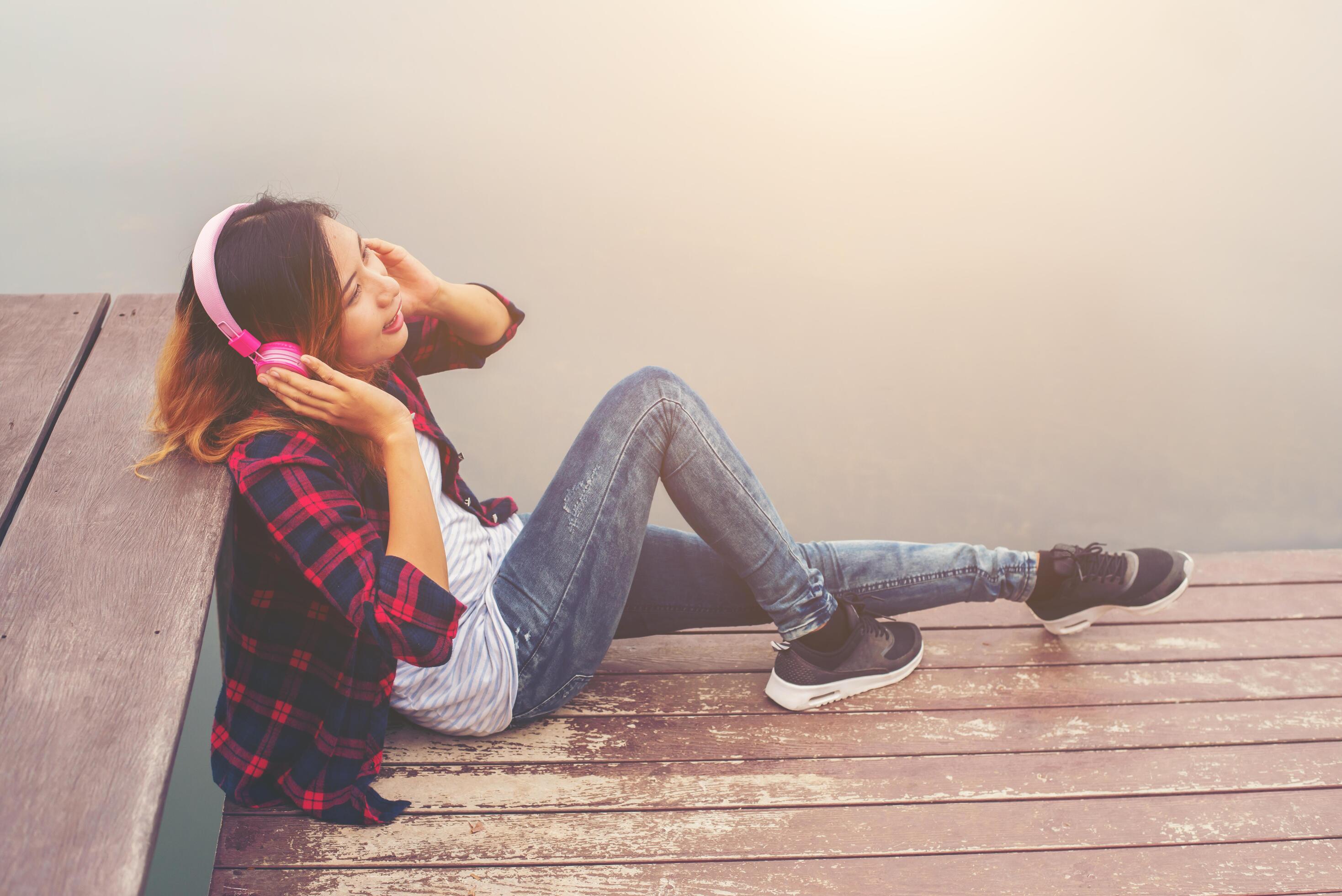 Portrait of young hipster woman with headphones sitting on pier and listening music relaxing with sunset,Lifestyle concept. Stock Free